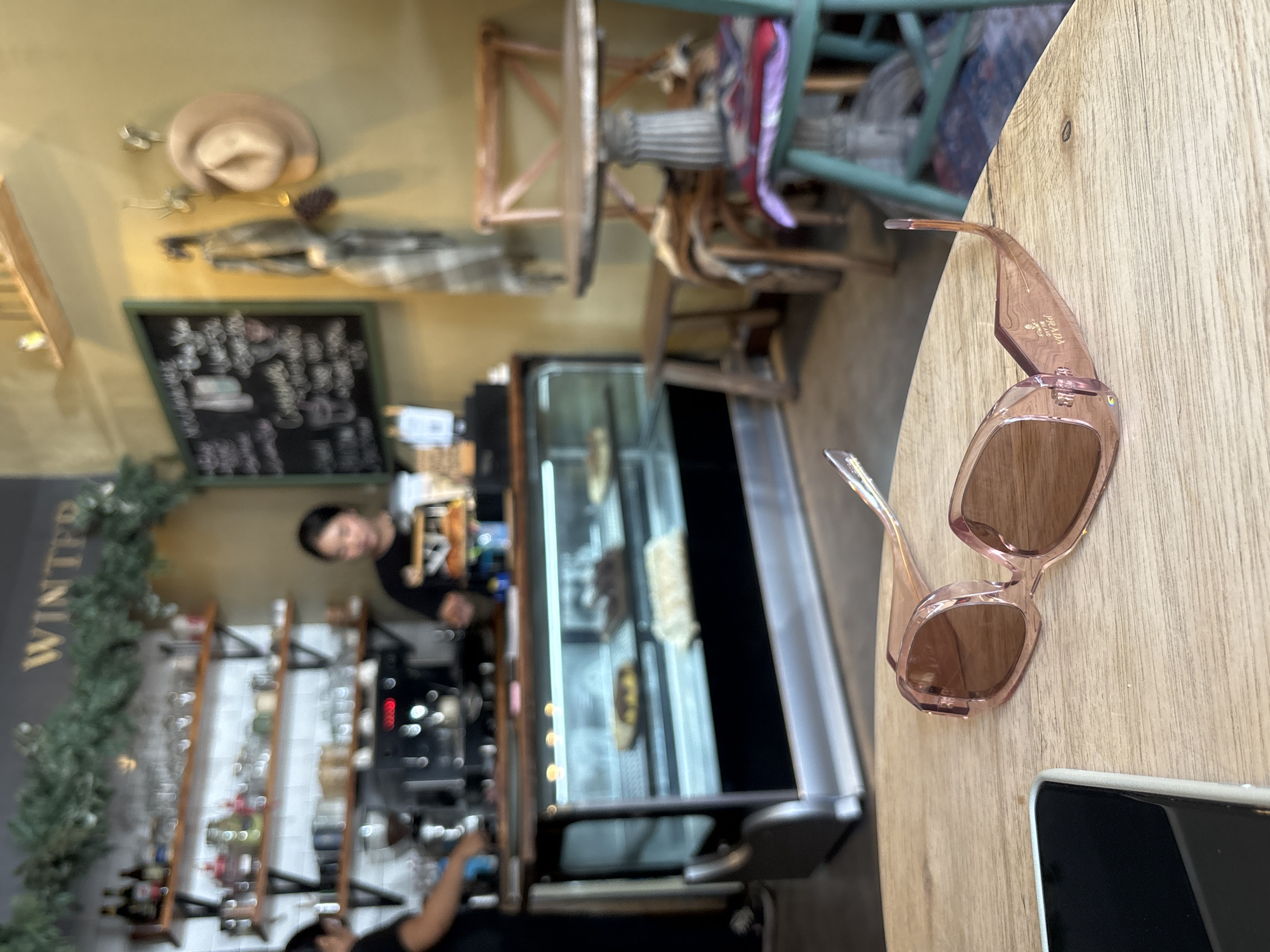 A pair of pink Prada sunglasses rest on a light wooden table in a café. A black smartphone lies next to the sunglasses. The background reveals the café's interior: a person in a black shirt stands behind a counter with an espresso machine. Behind the counter is a refrigerated display case filled with pastries. Shelves stocked with bottles line the wall on the left. A blackboard menu hangs above the counter. Greenery adorns the wall. A hat and coat are mounted on the wall above the blackboard. The scene is likely in a café in Kuta Selatan, Indonesia, as the café's interior design and product offerings appear to be consistent with the type of business expected in that location.