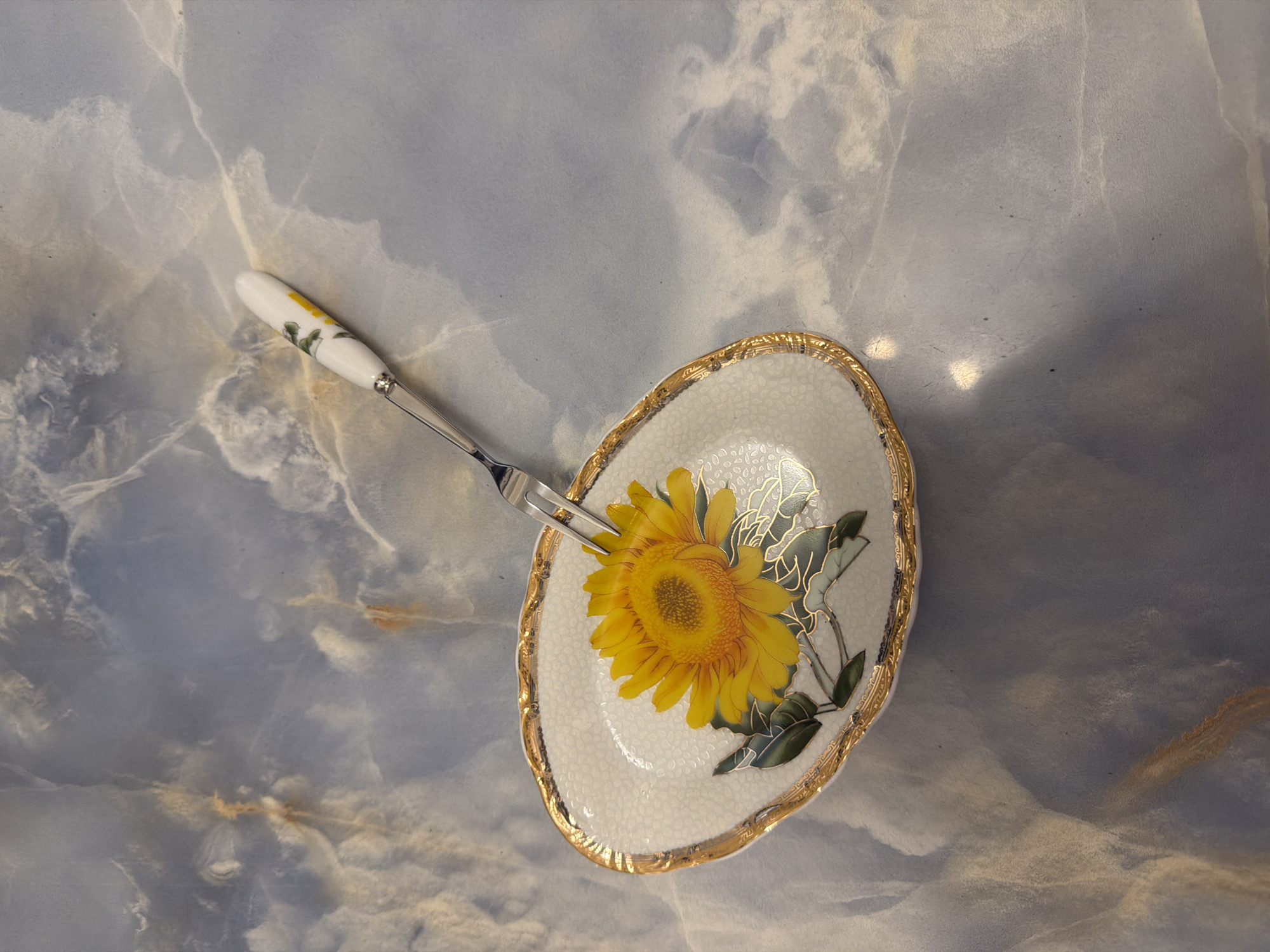 A metal fork with a white and yellow floral patterned handle rests diagonally across a small, oval, white ceramic dish decorated with a golden rim and a painted sunflower. The dish is set on a blue and grey marble-patterned surface. The lighting casts shadows and highlights on the surface and the objects. The scene is devoid of people and specific location indicators.