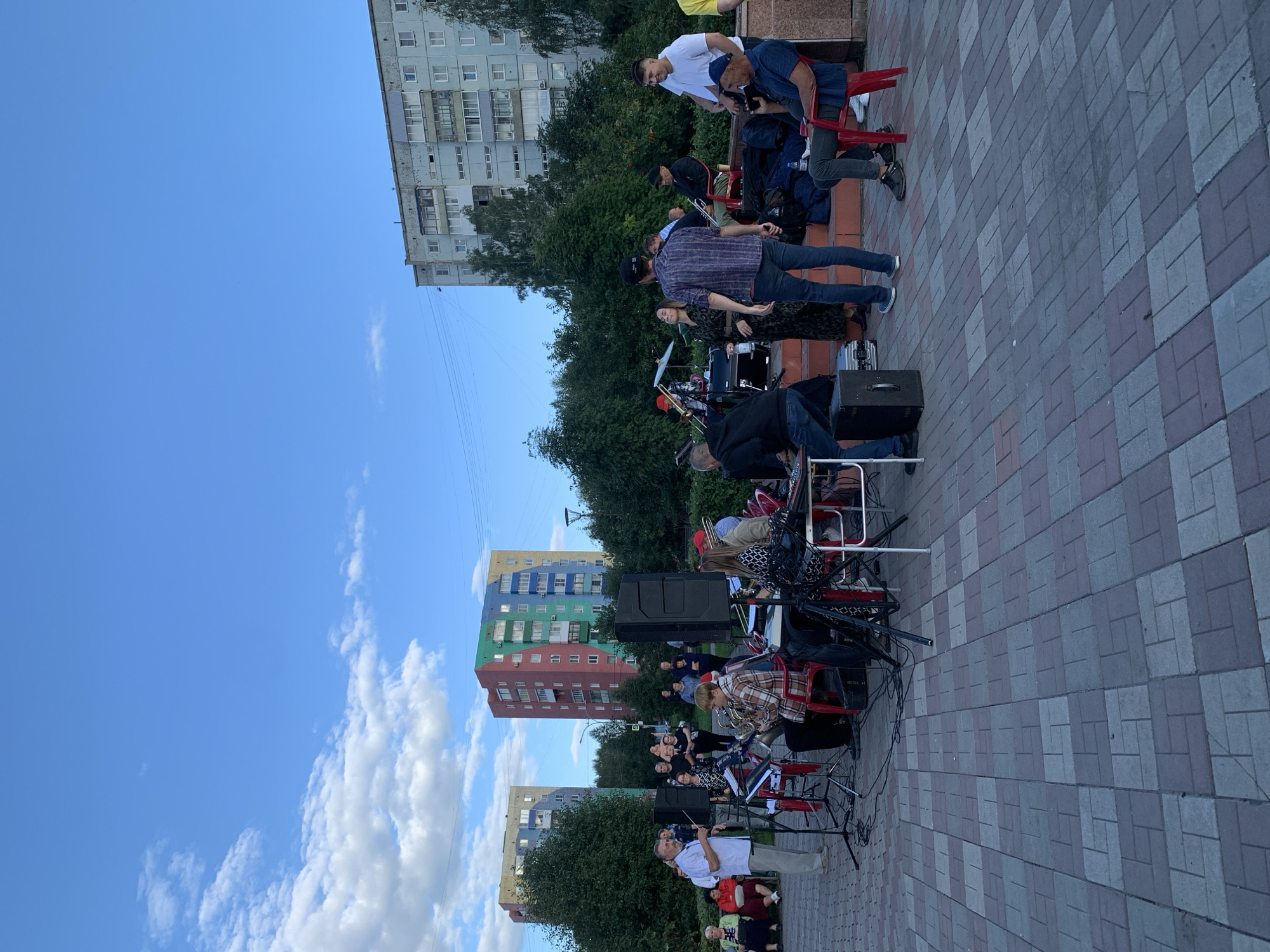 This image captures an outdoor public gathering, likely a concert or performance, in a city square in Prokopyevsk, Russia. The scene is set in front of several apartment buildings. In the foreground, a paved area with a patterned brick surface features a group of musicians and attendees. A band is set up with instruments, including a saxophone, keyboard, and drums, with several people actively playing or attending to equipment. Some attendees are seated on chairs, while others stand and watch.
Behind the musicians, a line of trees forms a backdrop. Further behind the trees, multi-story apartment buildings are visible. Two buildings in particular stand out: one is a grey, multi-story structure with balconies and windows, while another is notably colorful, painted in bold blocks of blue, green, yellow, red, and orange. The sky above is a clear blue with scattered white clouds, suggesting daytime. The overall atmosphere appears to be that of a community event taking place in an urban residential area.