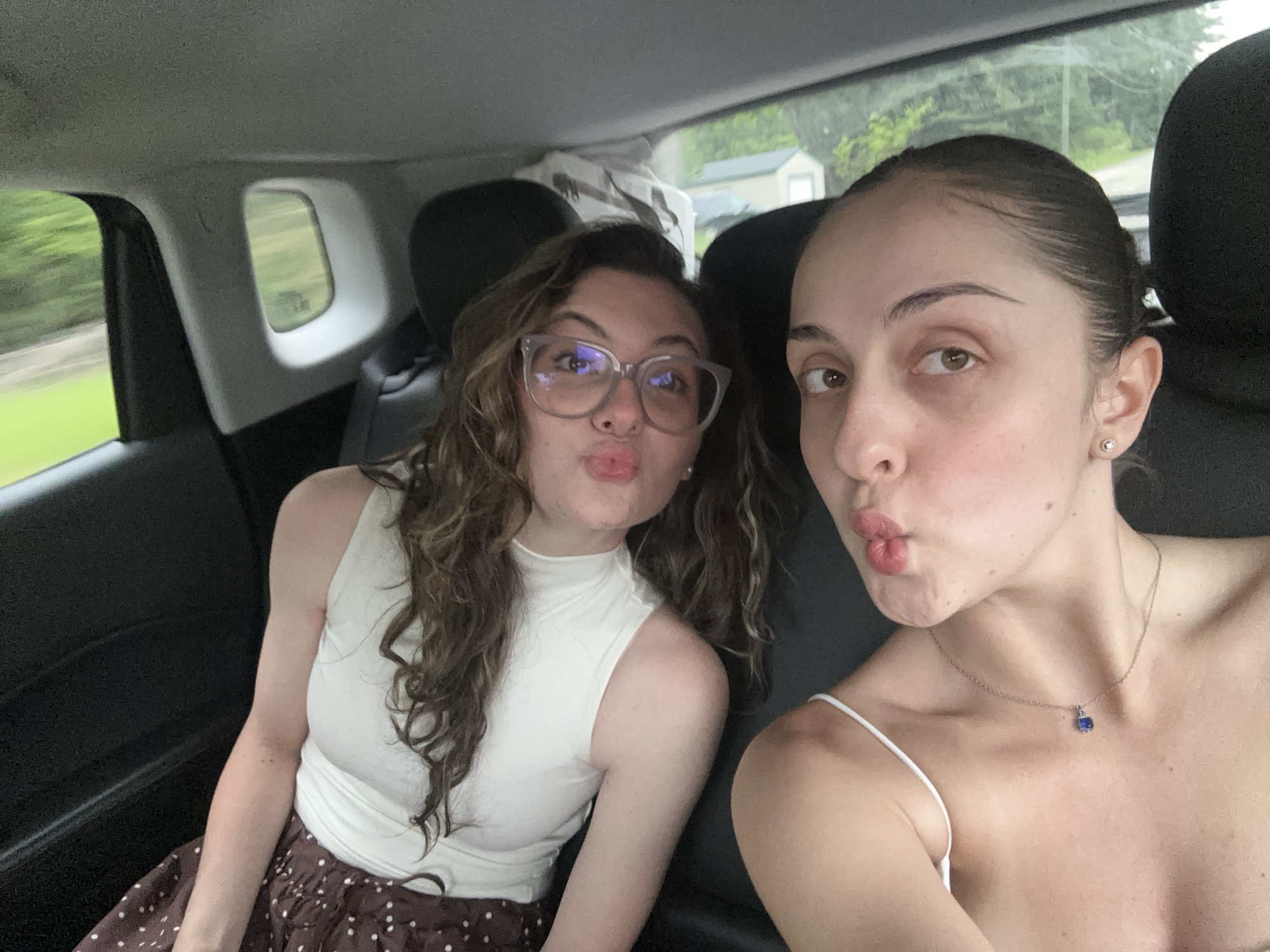 Two young women are seated in the back of a car. The woman on the left has curly brown hair and is wearing glasses, a white sleeveless top, and a brown and white polka-dotted skirt. She is puckering her lips. The woman on the right has dark hair pulled back and is wearing a white spaghetti strap top and a necklace with a blue pendant. She is also puckering her lips. The interior of the car is visible, including black seats and a window showing greenery outside. The scene appears to be a daytime photo taken while the car is in motion, likely on a road in or near Harbor Springs, Michigan, United States of America, given the residential building and trees visible through the window.