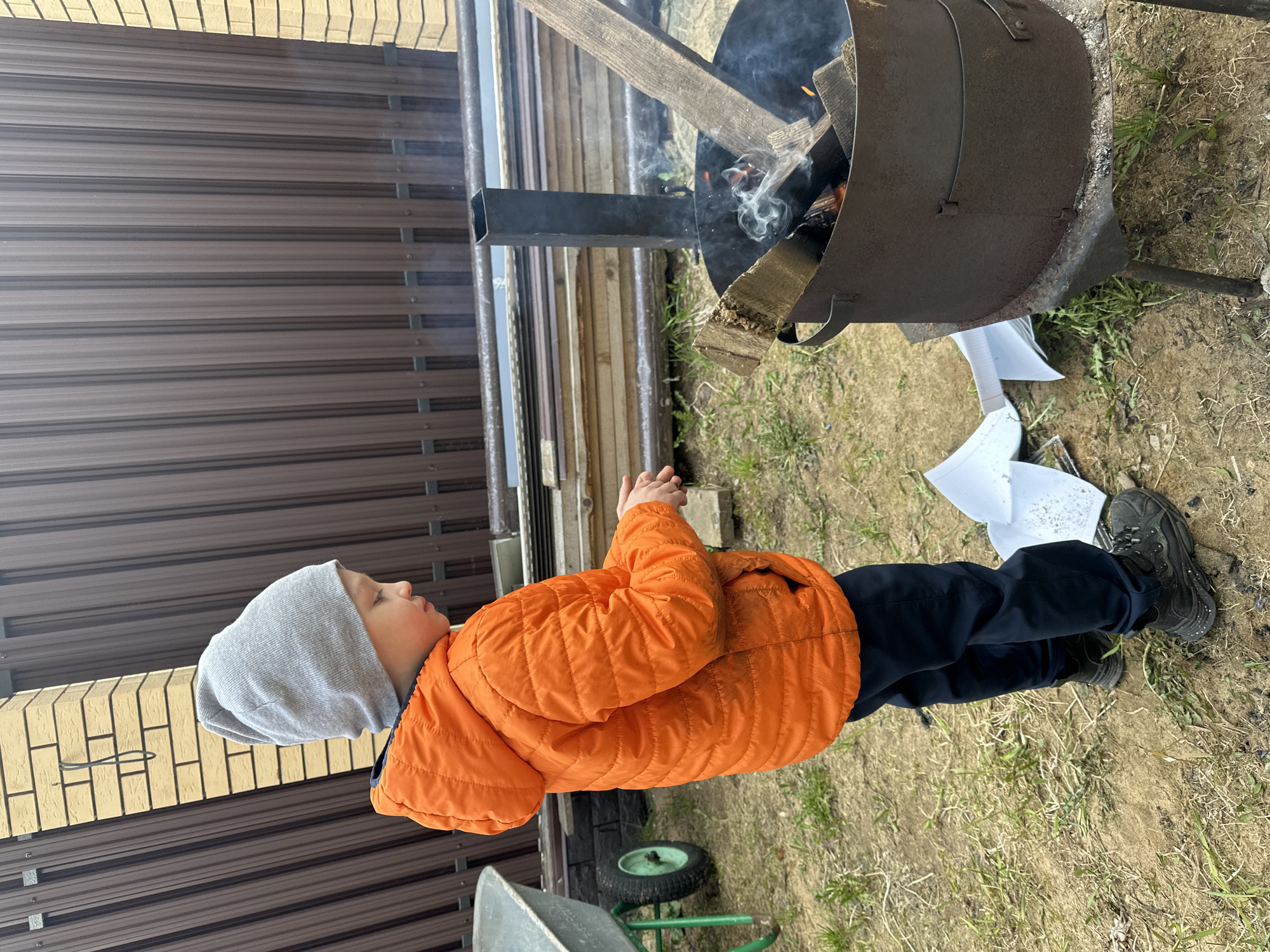 A child wearing an orange jacket and gray hat stands near a metal fire pit in слобода Старая, Russia. Wood and burning material are inside the pit, with smoke rising. Nearby is discarded white paper, and the ground is covered with grass and dirt. A metal fence and a brick structure are visible in the background.