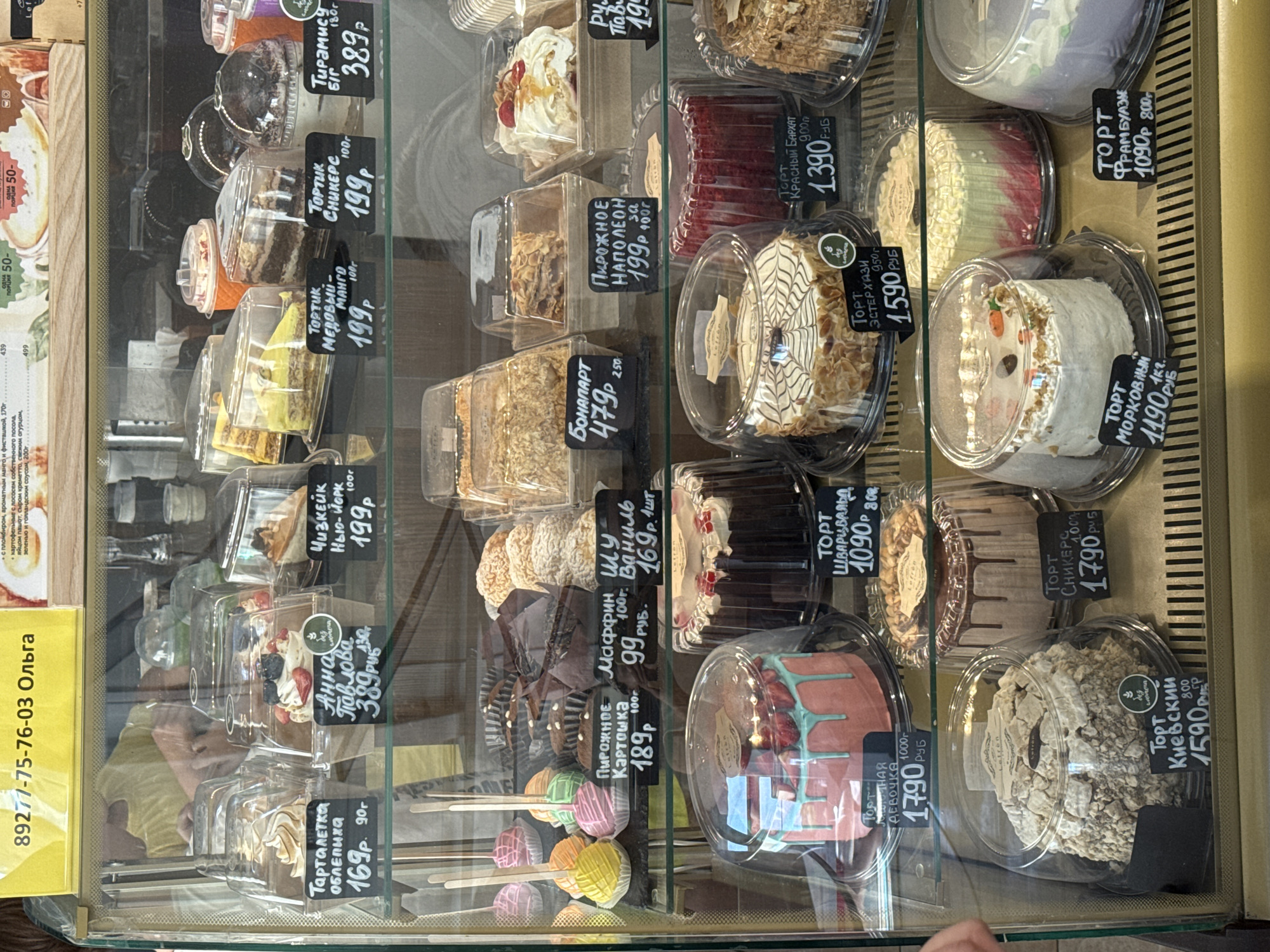 The image shows a refrigerated display case filled with various pastries and cakes. The display case is made of glass and metal, and it is well-lit, allowing for a clear view of the products. The pastries are arranged on multiple shelves, each presented in individual plastic containers.
On the top shelf, there are several individual servings of cakes and tarts. Some are topped with fruit and cream, while others appear to be layered or have intricate designs. Labels indicate different names like "Торт Анна Павлова" (Anna Pavlova Cake) and "Чизкейк Нью-Йорк" (New York Cheesecake) with prices and weights.
The middle shelf features a variety of smaller pastries, including what appear to be cupcakes or muffins, as well as some cake pops and larger cakes. Labels mention items like "Пирожное Картошка" (Potato Cake) and "Маффин Ваниль" (Vanilla Muffin).
The bottom shelf displays larger, whole cakes. One prominent cake is pink and blue with strawberries on top, labeled "Торт Нежная Девочка" (Tender Girl Cake). Another cake is dark chocolate with red detailing and is called "Торт Красный Бархат" (Red Velvet Cake). There are also cakes with elaborate frosting and toppings, such as one with a marbled design and another covered in what looks like nuts and meringue.
The background of the image is out of focus but suggests an indoor environment, possibly a bakery or a confectionery shop. A yellow sign with Cyrillic text, "89277-75-76-03 Ольга," is visible at the top of the display. The overall scene is one of abundance and variety in a commercial food setting.