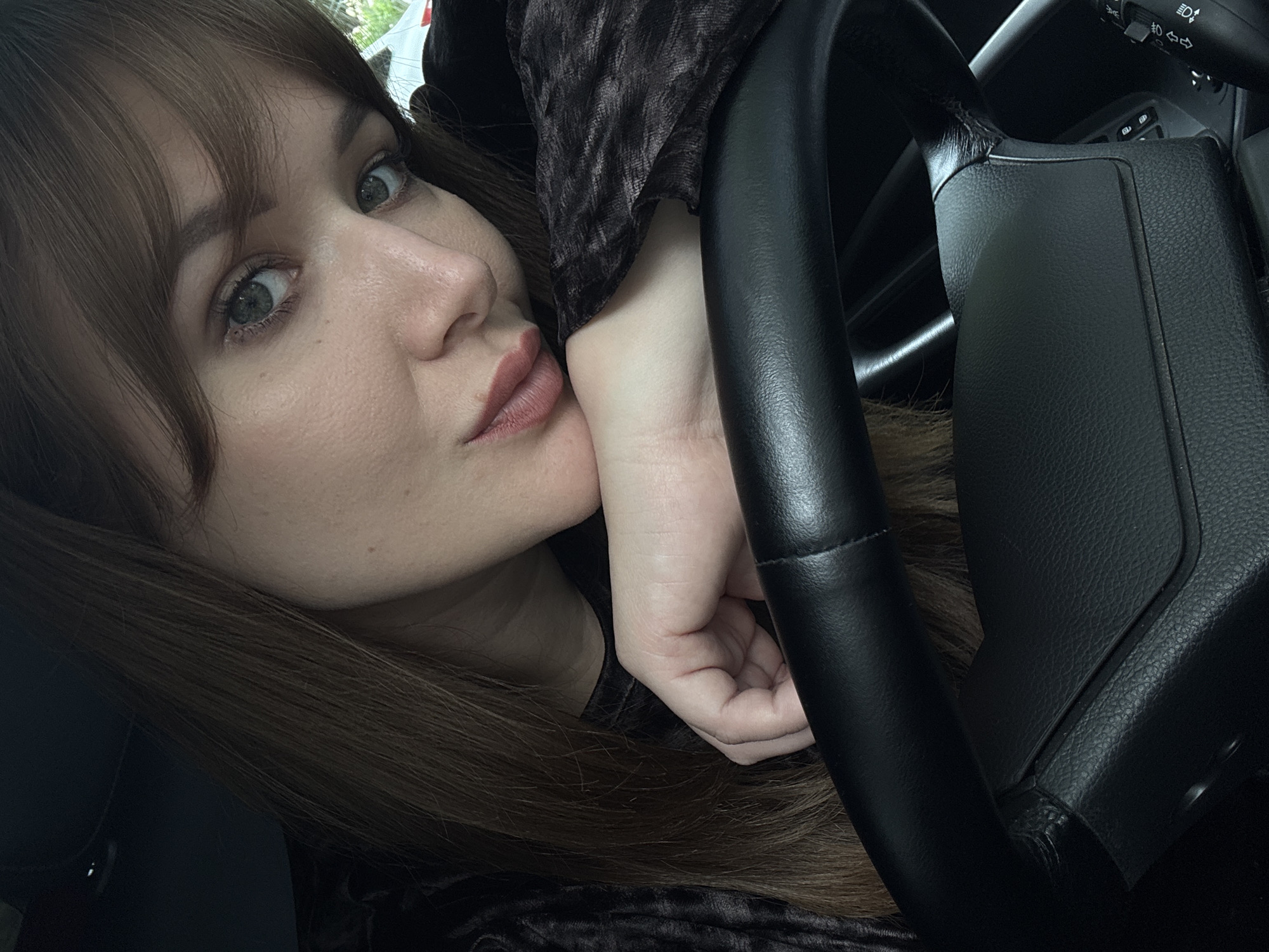 A close-up, slightly angled shot captures a woman leaning her head on her arm, which is resting on the steering wheel of a car. Her hair is brown and falls over her shoulder. She is wearing a dark, patterned top. The background is blurred, but suggests an indoor setting, possibly the interior of a car. The location is identified as Chaikovsky, Russia.