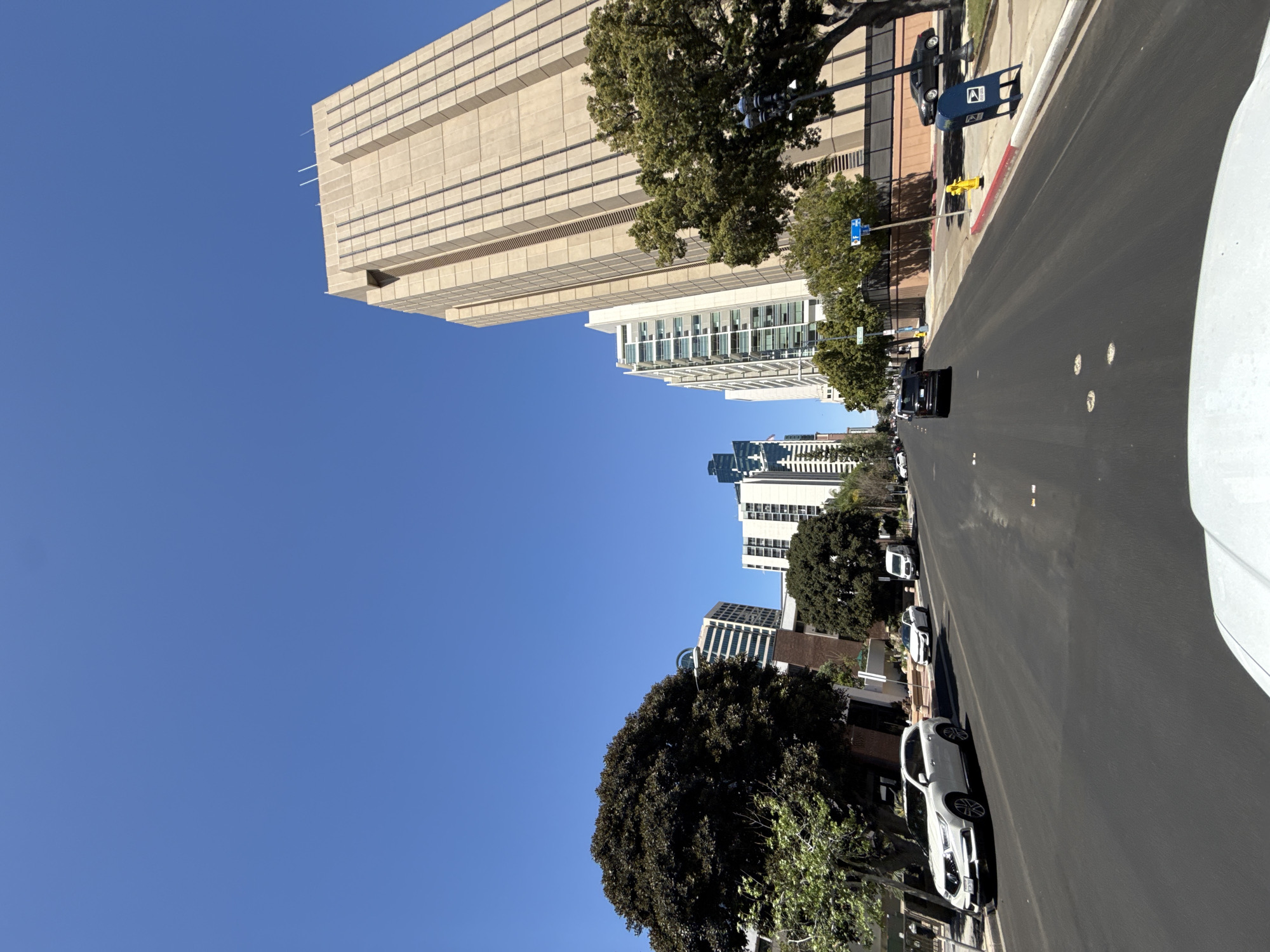 A street in San Diego, United States of America, flanked by a tall, beige-colored building on the left, stretches into the distance. Additional buildings line the street further down. Trees are interspersed between the buildings. A black sedan drives along the street, which has several white cars parked along the side of the road. A blue United States Postal Service mailbox is visible on the side of the street. The sky is a clear blue.