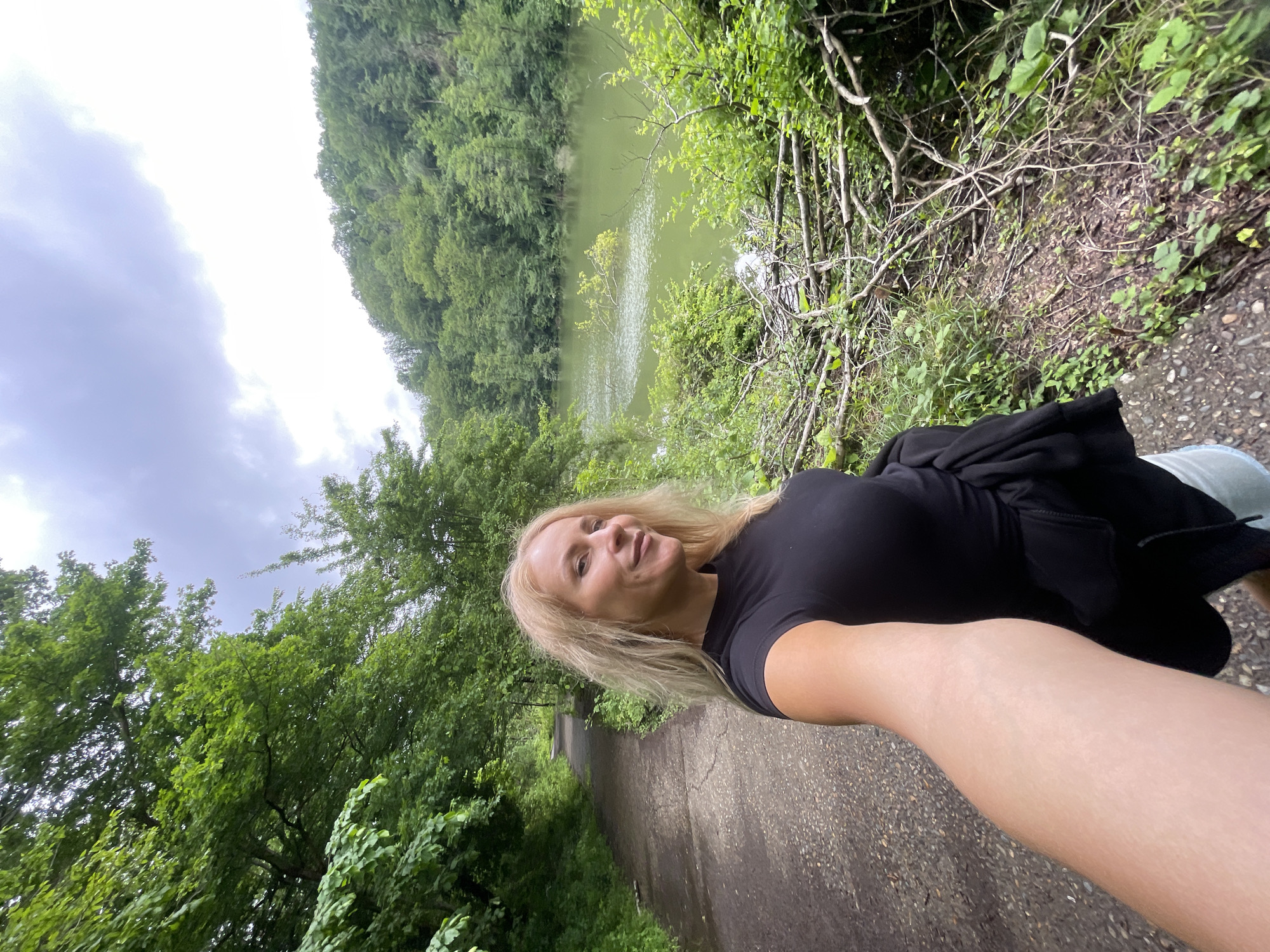 A woman with blonde hair is taking a selfie on a paved path. She is wearing a black t-shirt and has a black jacket slung over her shoulder. To her left is a dense, green forest. To her right, beyond some low-lying brush, is a body of water, possibly a lake. The sky is overcast. The path appears to be in a wooded area near Razdolnoye, Russia.