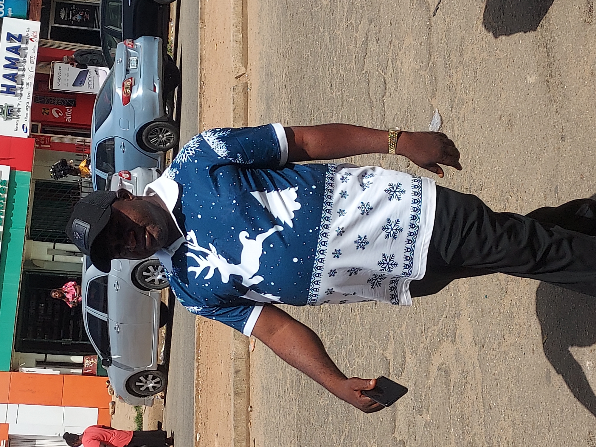 The image shows a man wearing a blue and white patterned shirt, black pants, and a black cap, standing on a street in Jos, Nigeria. He is holding a smartphone in his right hand and wearing a gold watch on his left wrist. The background includes buildings with storefronts, at least two cars and pedestrians. One of the storefronts has a sign that reads "HAMAZ" and advertises telecommunications services, including Airtel. Another appears to be a grocery store.