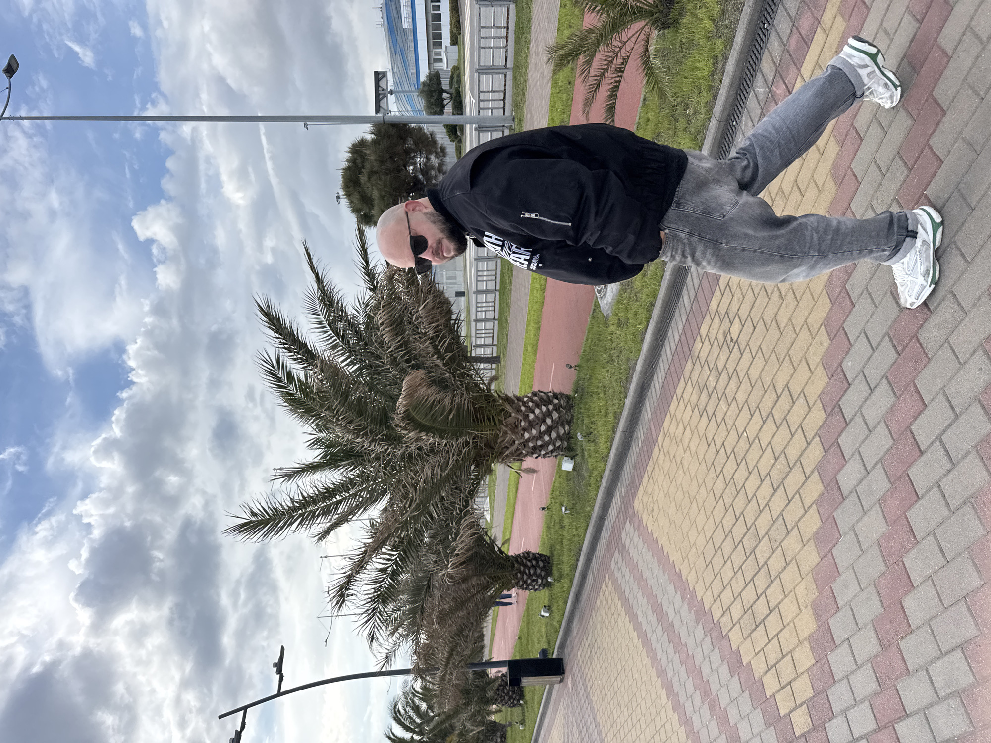 The image captures a man standing near palm trees on a brick-paved walkway in what appears to be a coastal area. The man is bald, wears sunglasses, a black jacket, gray jeans, and white sneakers with green accents. He is positioned on a walkway composed of interlocking rectangular bricks of yellow, gray, and pink hues. A line of palm trees extends into the distance alongside the walkway. Beyond the trees, a building with a blue roof and a glass facade can be seen. The sky is filled with numerous white and gray clouds. The location is the city of Sirius, Russia.