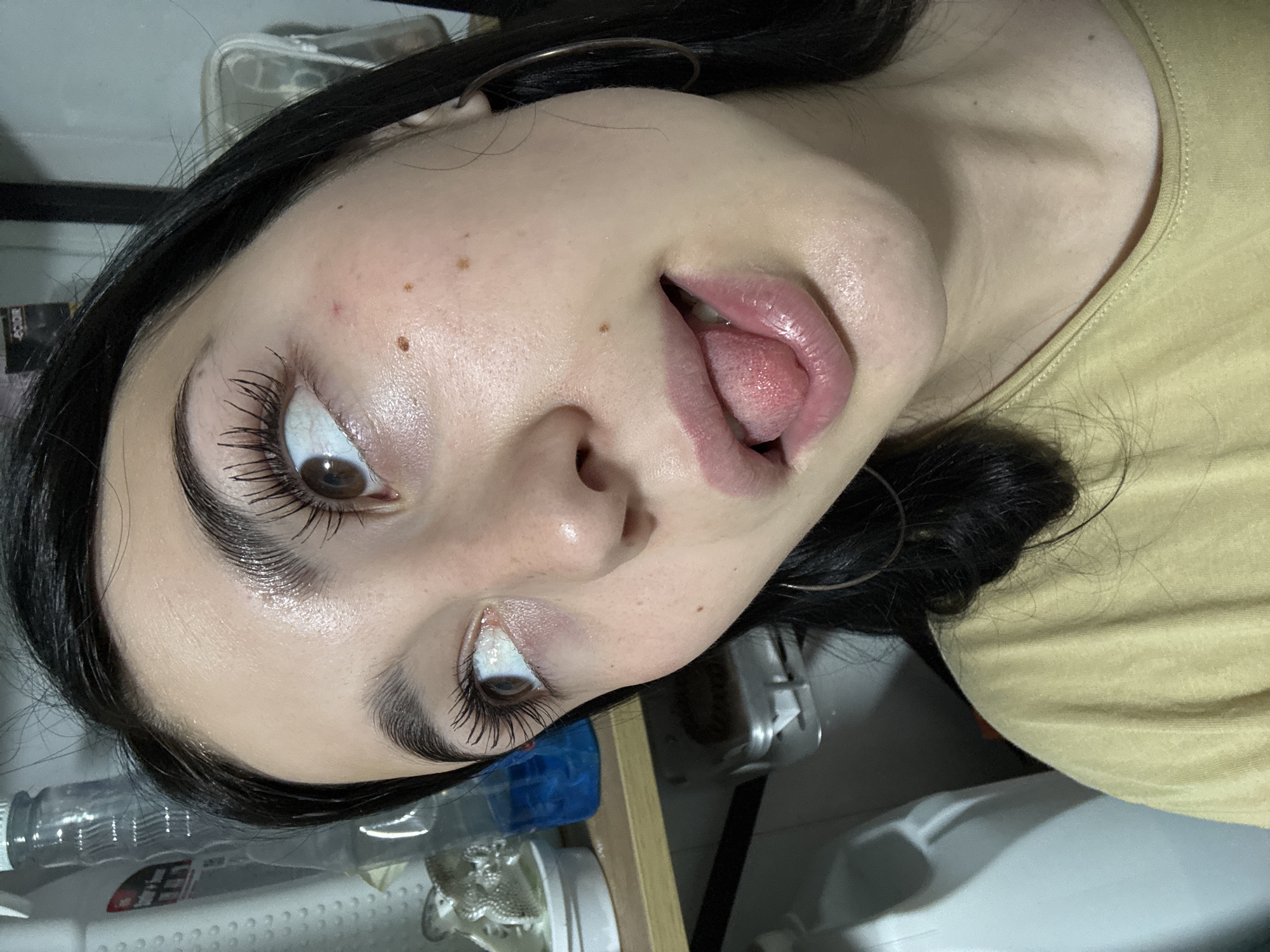 The image is a close-up, eye-level, medium shot of a young Caucasian female with dark hair and brown eyes. She is looking upwards and to the left, with her tongue slightly protruding from her mouth. Her lips are pink and glossy, and she has noticeable dark eyelashes and eyebrows. There are a few small freckles visible on her cheeks and bridge of her nose, and a small red mark above her left cheekbone. She is wearing a light yellow t-shirt and large hoop earrings.
The background is out of focus but appears to be an indoor setting with shelves. On the left, a clear plastic bottle with a white cap is visible, followed by a partially obscured blue bottle and a white container. On a shelf below, a metal object with a circular indentation can be seen. The overall lighting is soft.
The image does not contain any information about the location in the city of Фоки, Russia.