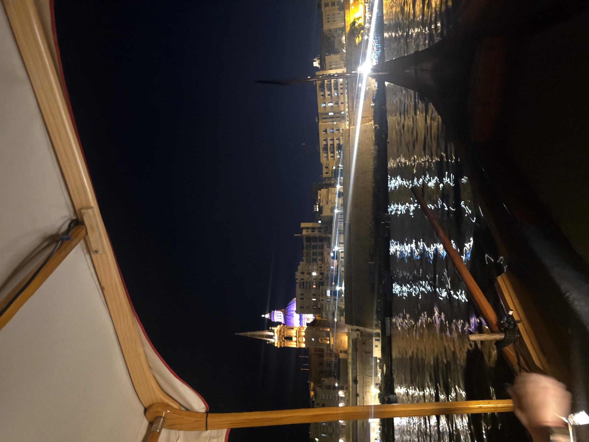 The image depicts a nighttime view of Valletta, Malta, from a moving boat, likely a gondola or similar vessel. In the foreground, the dark interior of the boat is visible, with a wooden oar resting across the frame. To the left, a person's hand and arm are blurred by motion, reaching towards the left edge of the image. Above, a canopy with wooden supports provides partial coverage.
The middle ground features the shimmering water of the Grand Harbour reflecting the lights of the city. Across the water, the historic architecture of Valletta is illuminated against the dark night sky. A prominent spire of a church, possibly St. Paul's Pro-Cathedral, is lit with warm yellow light, and a distinctive purple dome of another church, likely St. John's Co-Cathedral, is visible behind it. Buildings with illuminated windows line the waterfront, and bright streaks of light suggest other boat traffic or city lights. The overall atmosphere is one of a serene evening boat ride through the illuminated cityscape of Valletta.
