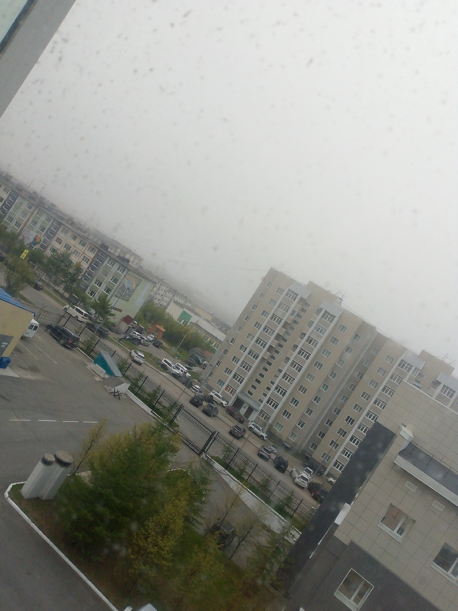 This is an image of a residential area in Magadan, Russia, taken from a high vantage point, likely a window, given the smudges and raindrops on the glass. The sky is overcast and foggy, contributing to a generally muted color palette. The scene depicts several multi-story apartment buildings, characteristic of Soviet-era urban planning, with light-colored facades and numerous windows. In the foreground, a paved area appears to be a street or parking lot, with several vehicles parked or driving. A black sedan is prominent in the lower left, and other cars, including white vans and SUVs, are scattered throughout. Green trees and some bushes are visible, suggesting a touch of nature integrated into the urban landscape. Fencing separates some of the properties. The overall impression is of a typical, somewhat dreary day in a Russian city, with the focus on residential architecture and urban infrastructure.