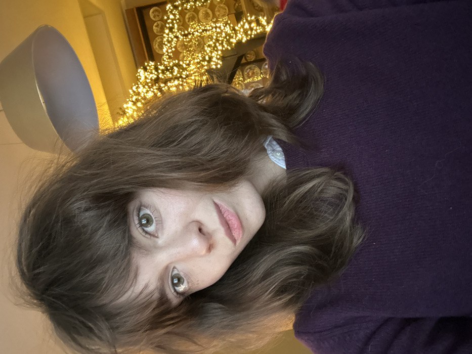 A photograph depicts an adult female positioned centrally, captured in a selfie-style composition within an interior setting in Tarasovka, Russia. The subject has medium brown, wavy hair with bangs and appears to have light-colored eyes, looking directly at the camera with a neutral expression. She is wearing a dark purple crew-neck sweater, with a light blue collared shirt visible underneath. The background is blurred, featuring a large, off-white circular pendant light fixture in the upper right. To the right and behind the subject, a significant portion of the background is illuminated by numerous warm-toned string lights, creating a densely lit, sparkling effect. Faint circular patterns, possibly decorative plates or wall hangings, are visible behind the string lights. The overall lighting is warm, emanating primarily from the decorative lights.
