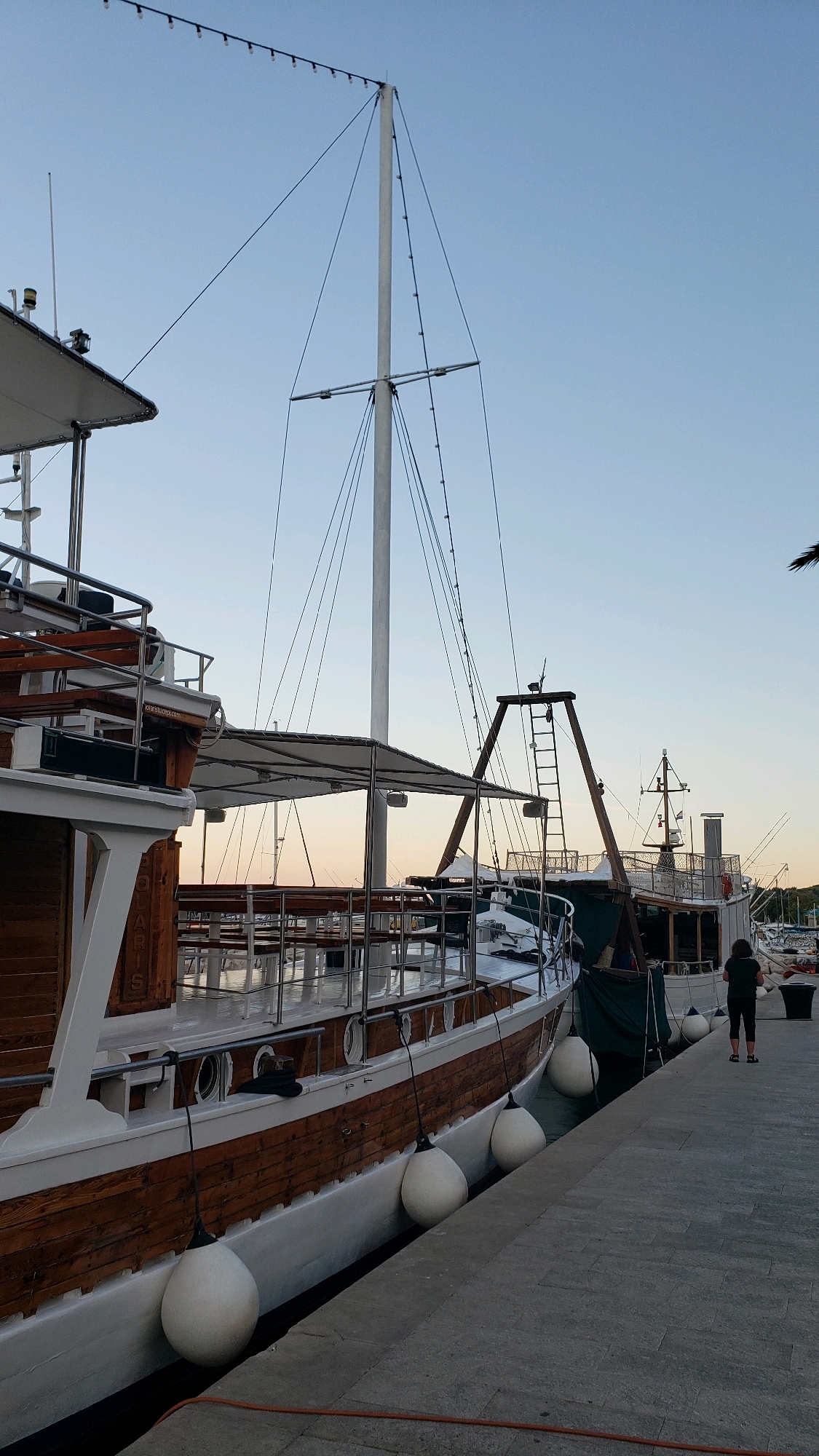 The image shows a docked boat in the marina of Općina Tučepi, Croatia. The boat is wooden with a white hull and is adorned with several large white buoys along its side. The sky is clear and blue, suggesting it is daytime. In the background, other boats and marina structures are visible. A person is standing on the dock facing away from the camera.
