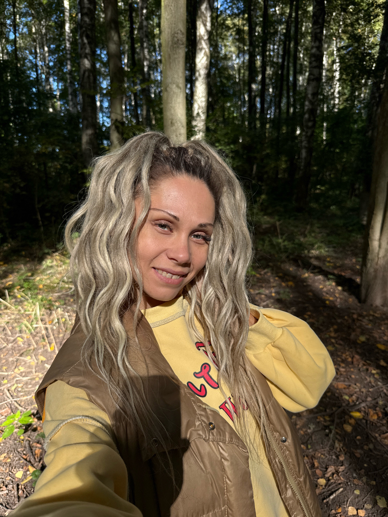 A woman with blonde, wavy hair tied in a high ponytail smiles directly at the camera. She is wearing a yellow sweatshirt with red lettering and a brown puffer vest. The background is a forest with tall trees and dappled sunlight on the ground. The location is the city of Rusanovo, Russia. The image appears to be a selfie taken outdoors.
