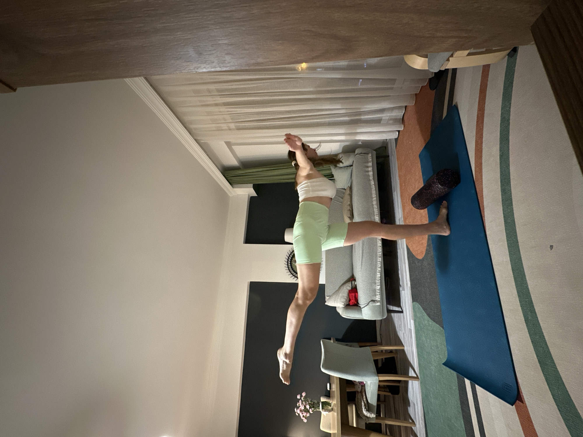 A woman wearing a white sports bra and light green athletic shorts is performing a yoga split in a room. She stands with her right foot on a blue yoga mat that is located on a carpet with tan, green, and orange stripes. A foam roller sits on the mat near her foot. A light grey couch is near the mat with a grey throw blanket. The background includes a dark grey wall and a beige wall. Curtains are to the side of the couch. A light-colored chair with a wooden back is near the dark grey wall with a wooden dining table underneath. A vase of pink flowers sits on the table. The room is located indoors. The city of Fethiye and the country of Turkey are not discernible from the information in the image.