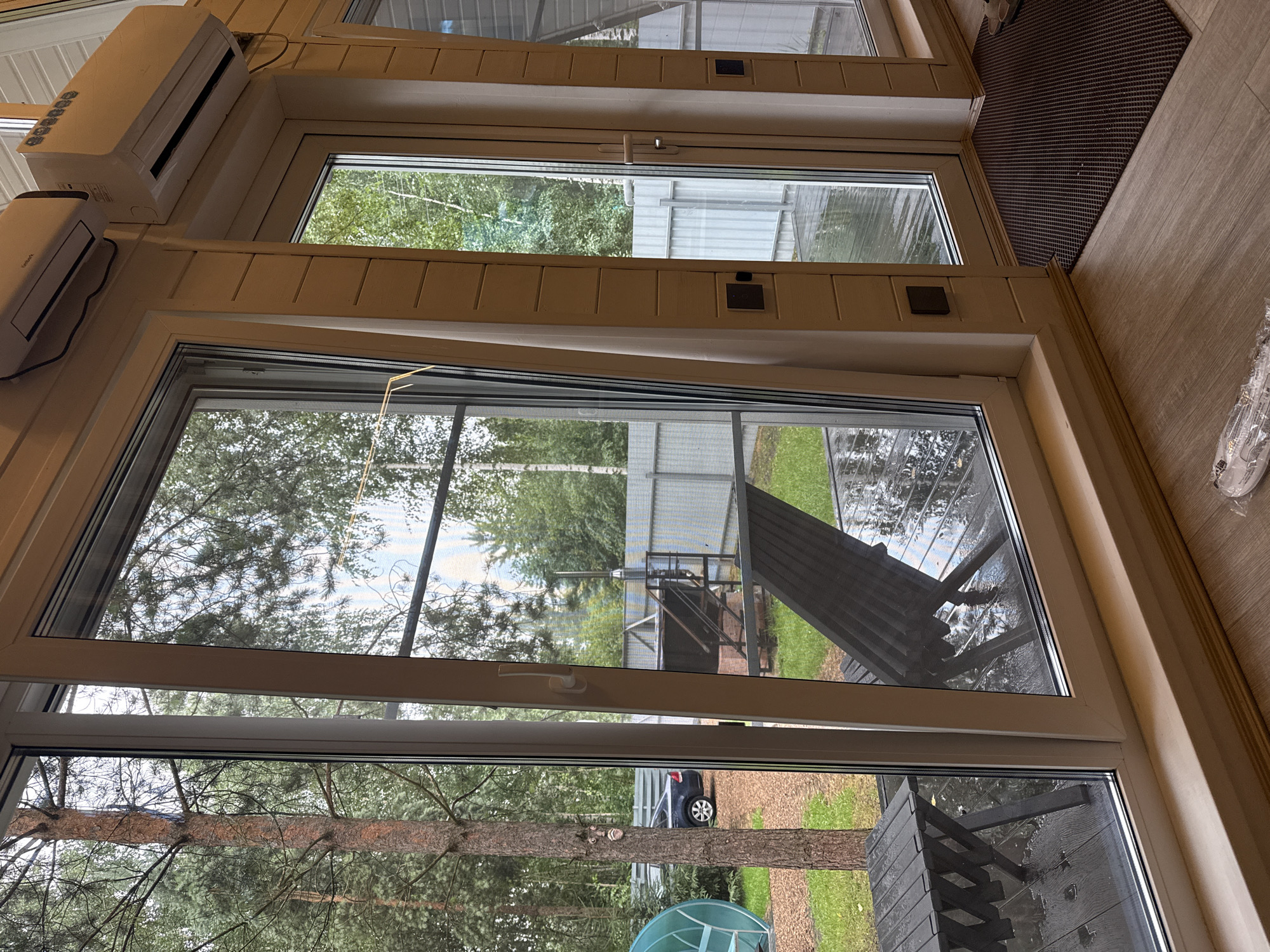 The image captures a view from inside a wooden-paneled room with large windows, looking out onto a wet outdoor area. The room has light-colored wooden walls and flooring. To the left, a large sliding glass door reveals a view of trees, a car, and some outdoor furniture. The outdoor patio appears to be constructed of dark wood planks that are wet, indicating recent rain. There are two dark-colored outdoor chairs and a table visible on the patio. In the background, a metal fence encloses the property, and beyond that, more trees and a building with a patterned facade can be seen. Two air conditioning units are mounted high on the interior wall above the windows. To the right, another large glass door leads outside, with a dark brown doormat on the floor in front of it. The overall scene suggests a domestic setting, possibly a dacha or country house, in the city of Prokudino, Russia.