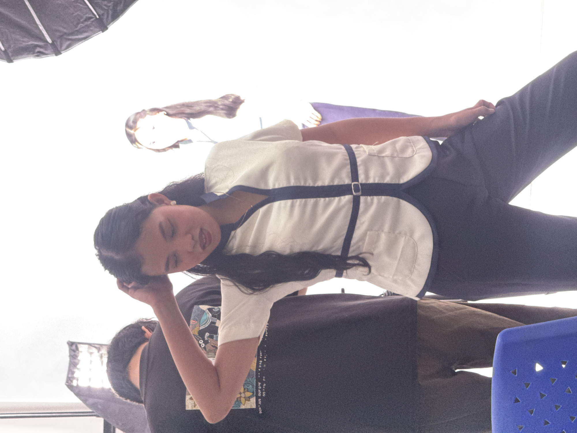 The image depicts a studio photoshoot in Davao City, Philippines. In the foreground, a young woman with dark hair and fair skin is posing, looking down with her right hand raised to her head. She is wearing a white short-sleeved top with blue trim and a matching belt, and dark pants. To her left, partially visible, is a person wearing a black t-shirt and brown pants, their back to the camera. In the background, another woman with long dark hair is standing, also looking downwards. The environment appears to be a brightly lit studio with photographic lighting equipment visible on the sides. A blue chair is partially visible in the bottom left corner. The overall scene suggests a professional or amateur photography session.