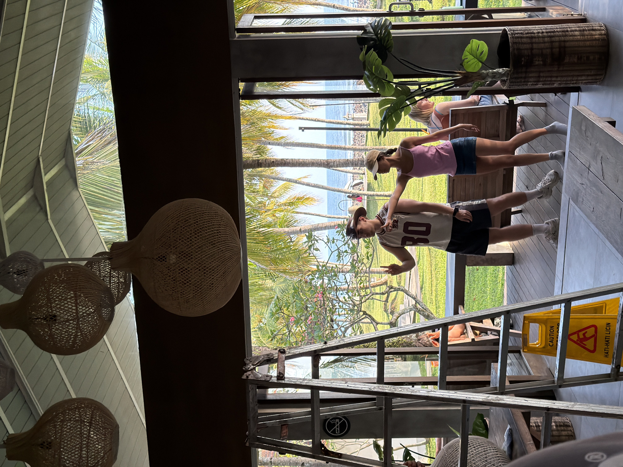 In Pekutatan, Indonesia, a man wearing a white tank top with the number 30 and dark shorts, and a woman in a pink tank top and denim skirt, both wearing hats, stand on a wooden deck. The man is making a peace sign with his left hand and pointing with his right. The woman is pointing to her right. Behind them, palm trees and the ocean are visible through an open doorway. A stepladder is positioned to the left of the couple, with a yellow caution sign at its base that reads "CAUTION WET FLOOR" and "HATI-HATI LICIN." Several large, woven pendant lamps hang from the ceiling above.
