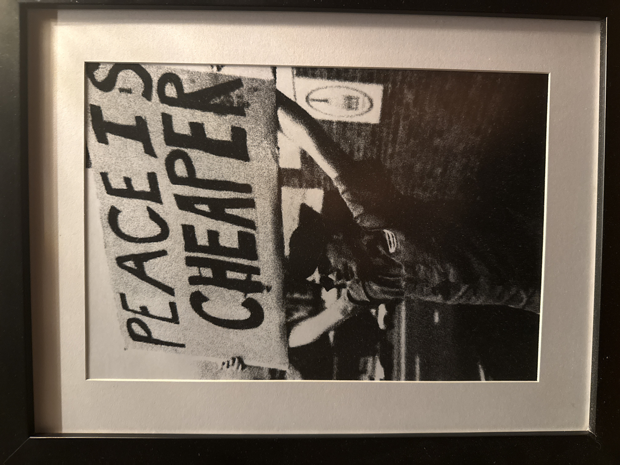 A framed black and white photograph of a protest sign that reads "PEACE IS CHEAPER". The sign is held by people whose faces are obscured, but there is a man in a hat behind the sign. The photo is framed with a white mat and a black frame. There is no indication this image is from North Kuta, Indonesia.