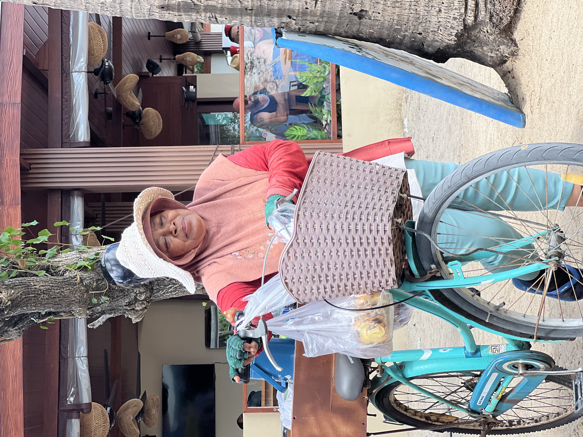 In Kuta, Indonesia, a woman wearing a pink hijab and a light-colored sun hat stands beside a turquoise bicycle. She is dressed in a coral-colored shirt and light blue pants, with green fingerless gloves on her hands. The bicycle has a woven basket attached to the handlebars, holding plastic bags filled with food items. A wooden cart is positioned behind the bicycle. In the background, a restaurant with rattan light fixtures and ceiling fans is visible, and through a glass window, two men are seen inside. A palm tree trunk and a blue surfboard lean against it. The ground is sandy, indicating an outdoor, possibly beachside, setting.