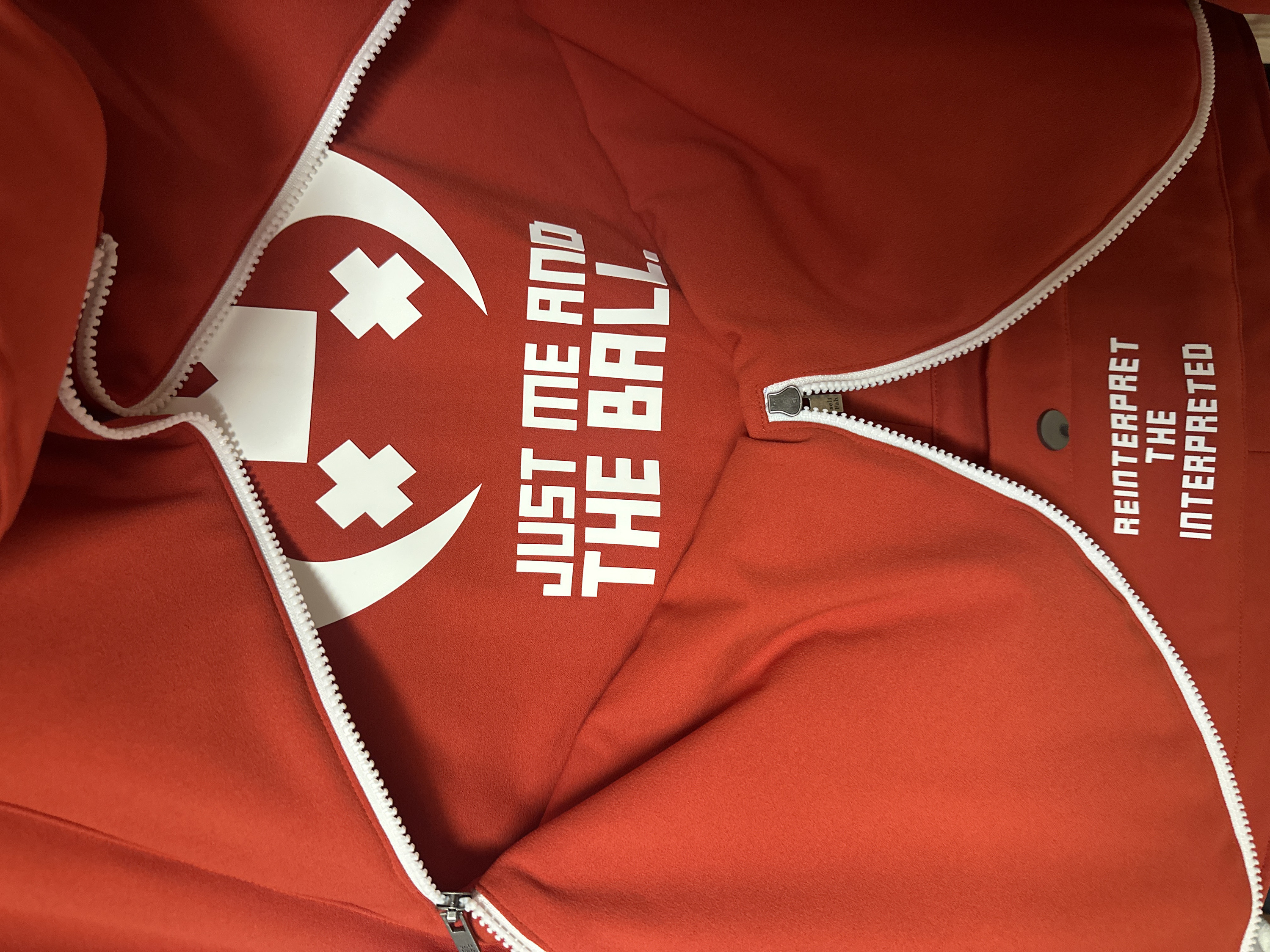 The image shows a red jacket or hoodie laid flat, viewed from above, with its central zipper partially open. The jacket is made of a soft, possibly fleece-like fabric, displaying subtle textures and natural folds.
A prominent white plastic zipper runs diagonally across the image from the top-right to the bottom-left, revealing the interior of the garment. The zipper pull is metallic and silver-colored, bearing a faint inscription that appears to be "YKK".
On the left side of the jacket (when viewed as if the jacket were upright), a large white graphic is visible. This graphic consists of a broad, upward-curving crescent shape at the top, and two distinct 'X' shapes positioned below it, collectively forming a stylized, smiling face or character. Adjacent to this graphic, to its right, is a block of white text in an uppercase, sans-serif, blocky font. The text reads: "JUST ME AND THE BALL", though the final letters "LL" of "BALL" are partially obscured or cut off at the edge of the image.
On the right side of the jacket, positioned higher up near what would be the collar area when worn, is another set of white, uppercase text. This text is arranged vertically on the jacket's design but appears horizontally oriented due to the image's rotation. It reads: "REINTERPRET THE INTERPRETED", with each word stacked below the previous one.
The dominant color of the jacket is a deep red, which provides a strong, eye-catching contrast with all the white elements—the zipper, the graphic, and the text. The lighting is even, suggesting an indoor setting. No specific activity is occurring; the garment is simply displayed.