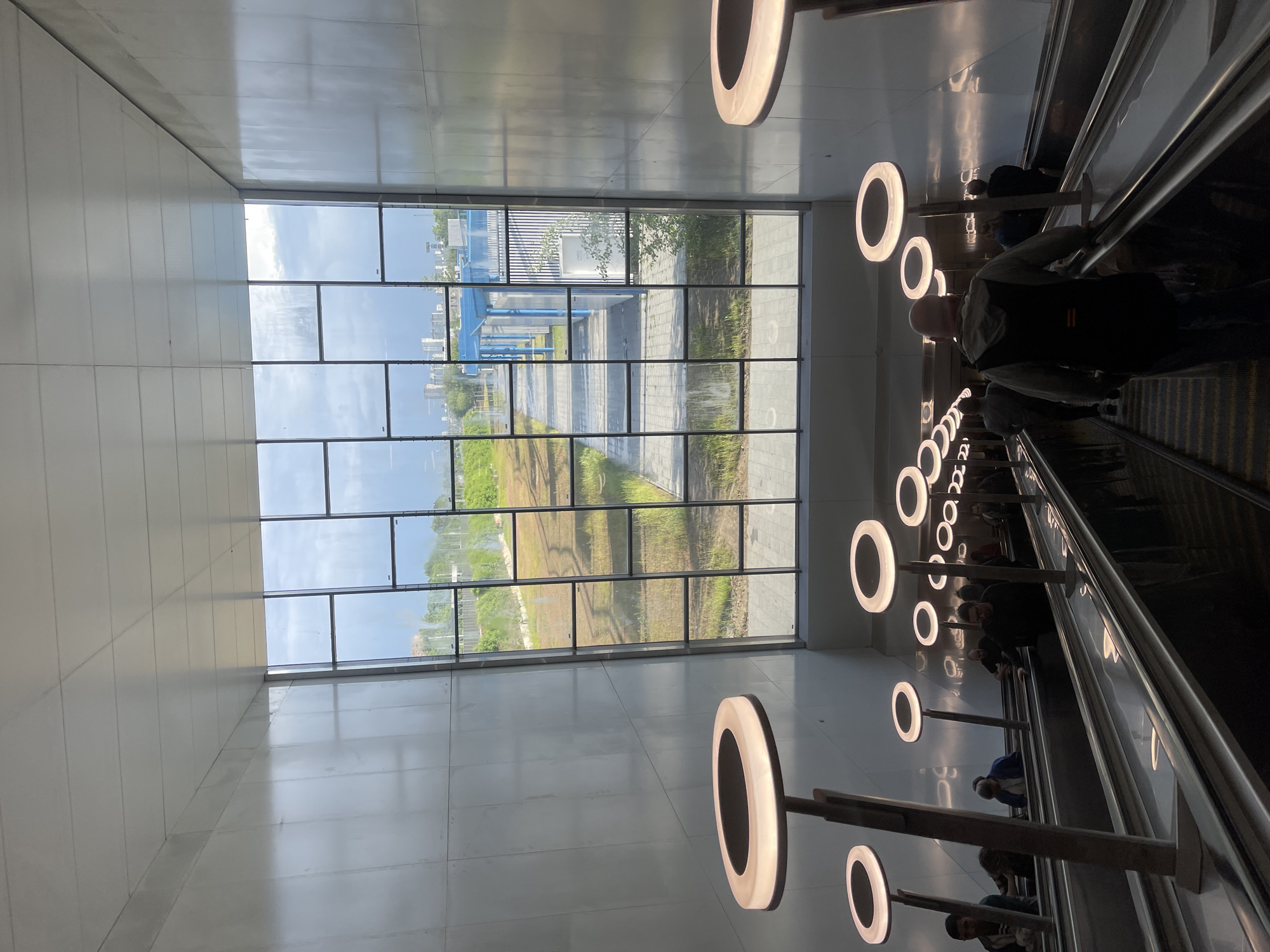 Here is a detailed description of the image:
The image captures an indoor scene, seemingly inside a modern transportation hub. The perspective is angled, emphasizing the depth of the space.
A large window comprised of multiple panes dominates the left side of the image, offering a view of an exterior landscape. It appears to be an open area with grass and foliage, and what might be a pedestrian pathway or construction site. Buildings are visible in the distance. The sky above is partly cloudy.
To the right, an escalator ascends, carrying multiple people. The individuals on the escalator are dressed in casual clothing, and their faces are mostly indistinct due to the distance and lighting.
Circular, ring-shaped light fixtures are mounted on poles along the side of the escalator, illuminating the scene. The walls and surfaces are primarily white and glossy, reflecting light and enhancing the brightness of the space.