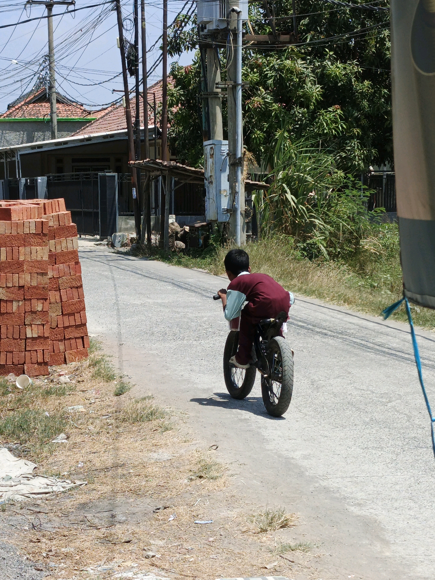 The image depicts a child riding a small motorcycle on a paved road in Santing, Indonesia. The child, viewed from behind, wears a maroon shirt with light blue sleeves. The motorcycle appears to be a child-sized dirt bike. To the left of the road, a stack of red bricks is piled high. In the background, houses with tiled roofs and abundant green foliage are visible, along with numerous utility poles and electrical wires. The environment suggests a residential area with construction materials present. The lighting indicates it is daytime with a clear sky.