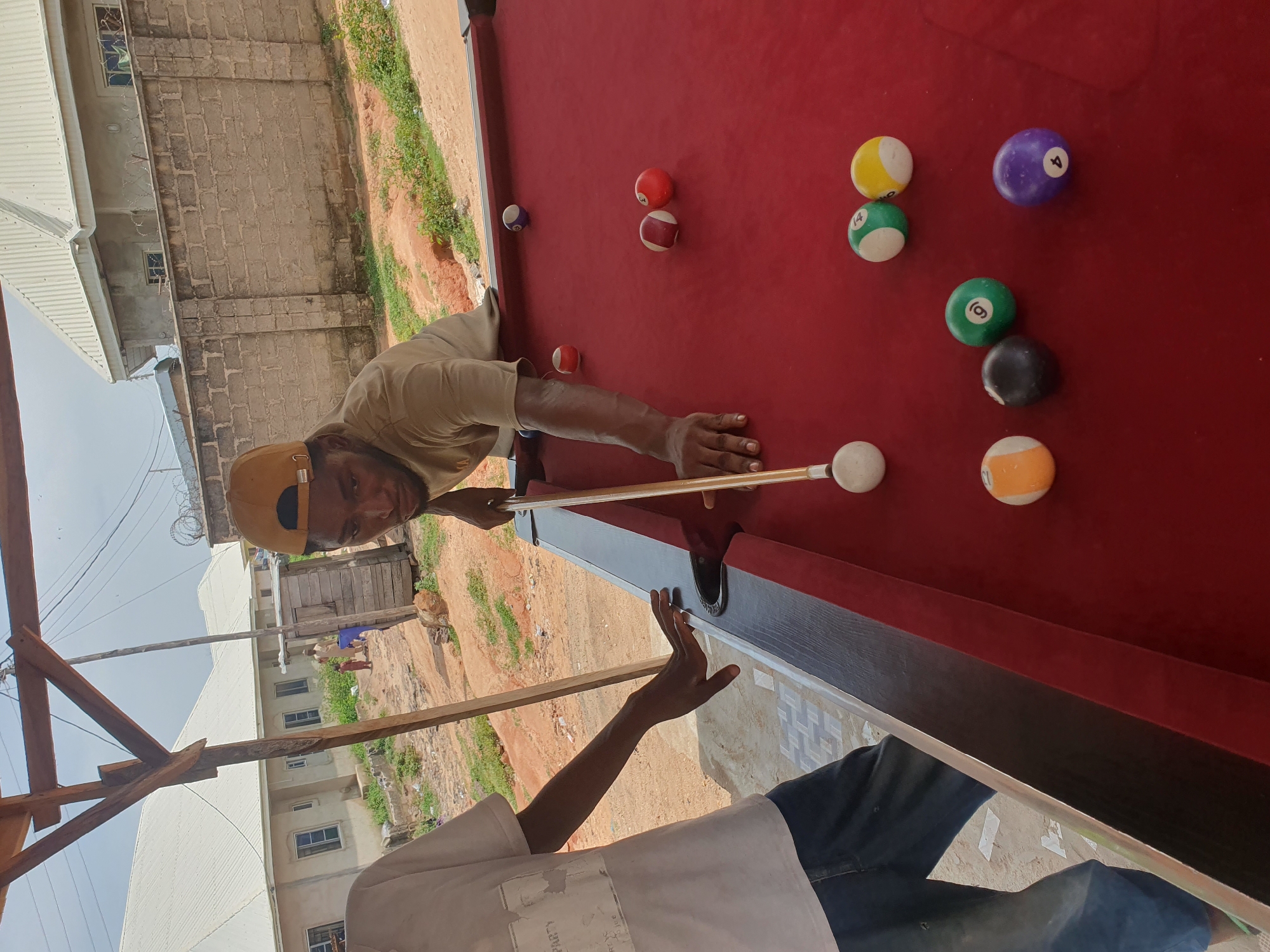 This is a medium-angle, eye-level, outdoor, full shot of two men playing pool at a billiard table. The billiard table has a red felt surface. In the foreground, the left side of a man's torso and legs are visible; he is wearing a white t-shirt with an abstract grey design and blue jeans. His left hand is resting on the edge of the billiard table. To the right, a man wearing a tan t-shirt and a yellow baseball cap is leaning over the billiard table, holding a pool cue. He is in the process of striking a white cue ball. Several billiard balls are scattered across the table, including a solid red ball, a red and white striped ball, a solid blue ball, a solid green ball with the number 9, a solid yellow ball, a solid black ball, a solid purple ball with the number 8, and a solid orange ball with the number 11. The background shows a dirt-covered ground, some overgrown grass, a low brick wall, and several residential buildings with white corrugated metal roofs. The scene appears to be in Lokoja, Nigeria.