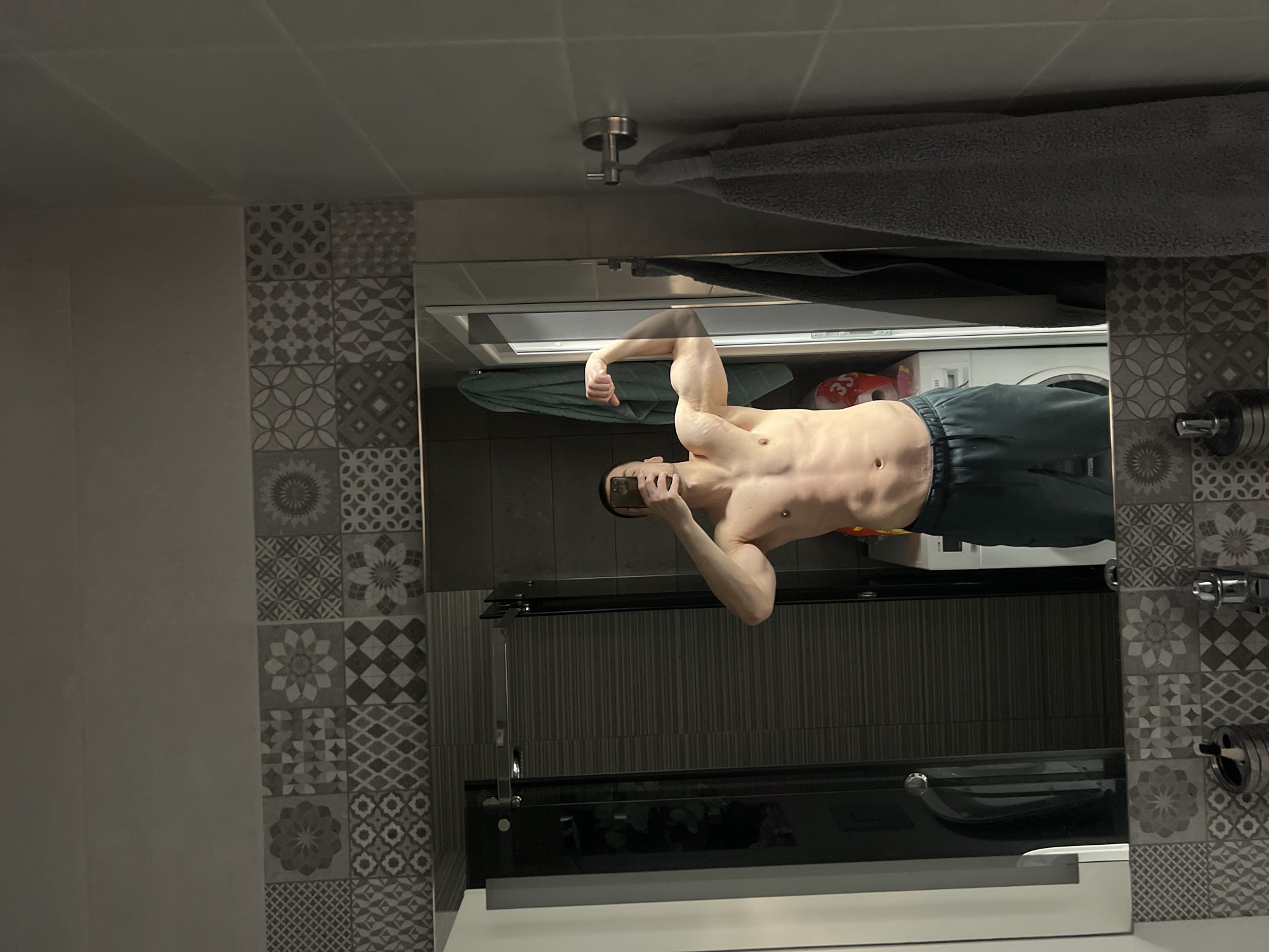 A shirtless man is reflected in a bathroom mirror, flexing his bicep while holding a phone to take a picture. He is wearing dark grey sweatpants. A washing machine and laundry supplies are visible in the background. The bathroom has patterned grey and white tiles. There is a towel hanging above the mirror. The specific location in Burgas, Bulgaria, cannot be determined from the given visual information.