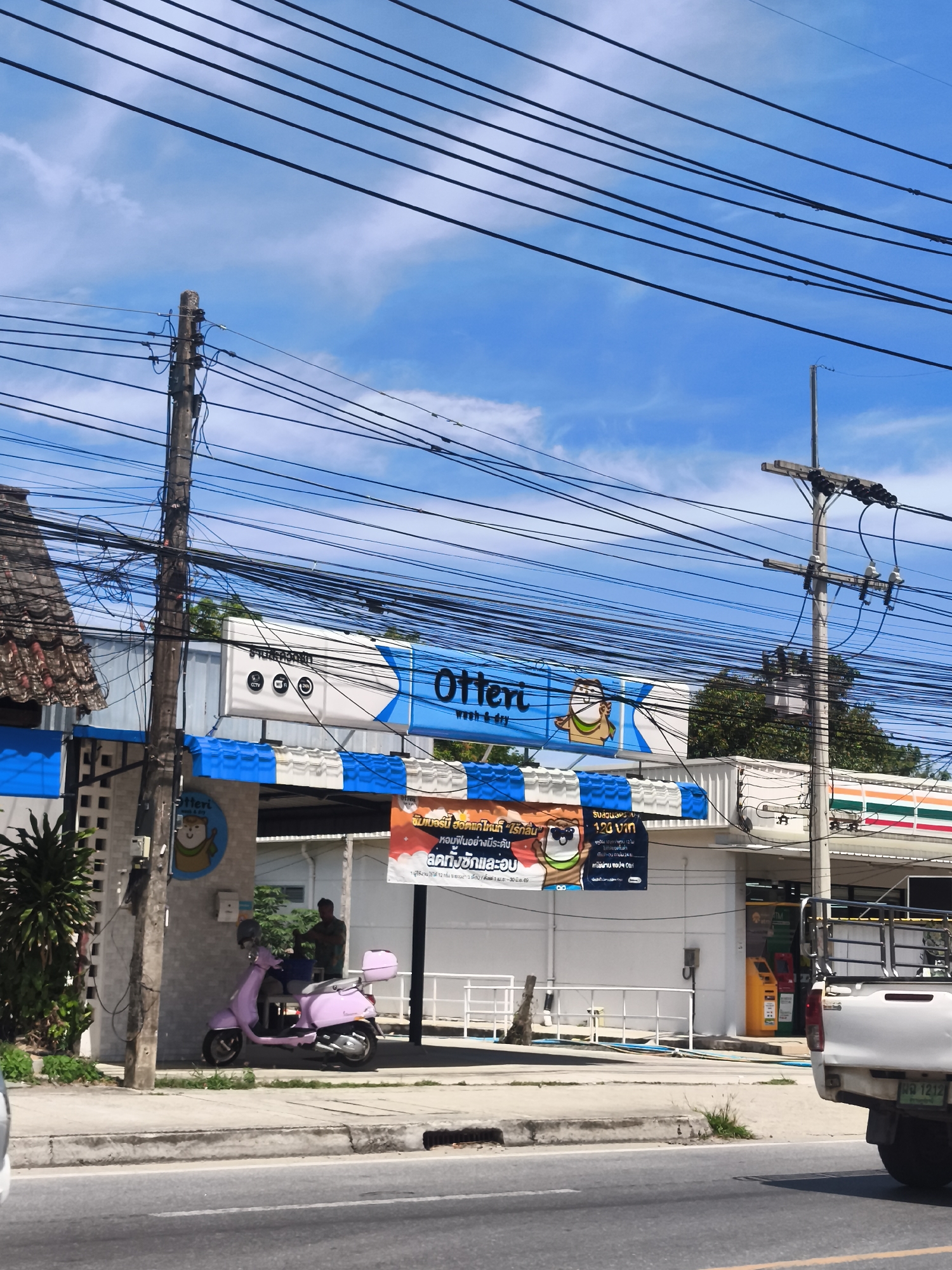 A street view captures a commercial establishment identified by a prominent blue and white awning with the word "Otten" and a cartoon hamster wearing glasses. The establishment appears to be a laundry or dry-cleaning service, as indicated by icons for CCTV, WiFi, and possibly another service related to washing. A pink Vespa scooter is parked on the sidewalk in front of the building. A man is standing near the scooter, looking towards the establishment. Next to the building, a 7-Eleven convenience store is visible. The scene is characterized by an abundance of overhead electrical wires and utility poles, suggesting an urban or semi-urban setting in Baan Hua Thanon, Thailand. A white pickup truck is parked in the foreground, partially obscuring the view of the building. The sky is blue with scattered white clouds.