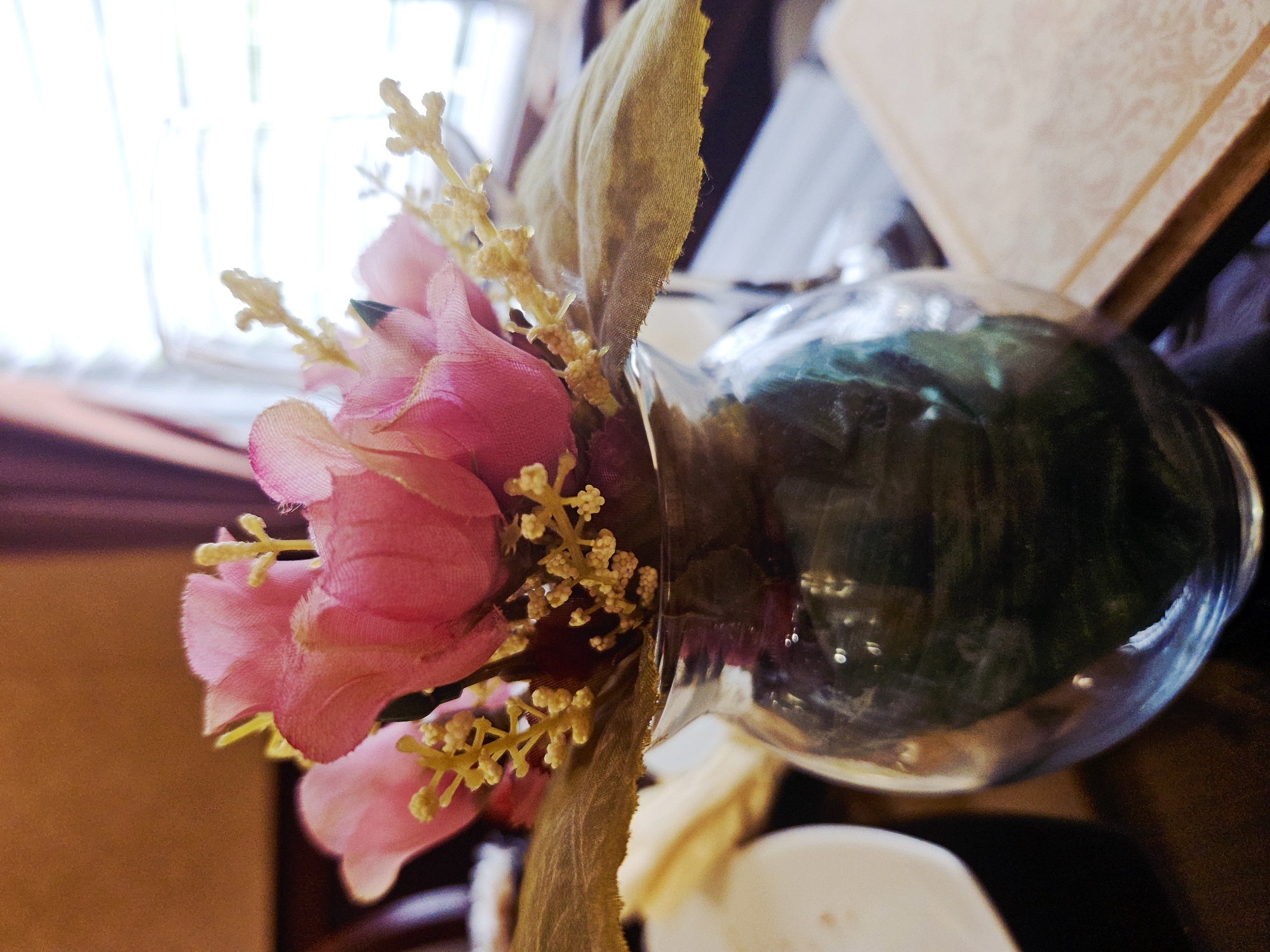 A close-up, eye-level shot showcases a clear glass vase holding a small arrangement of artificial pink flowers and dried foliage. The vase, which appears to be filled with dark liquid, is placed on a dark, reflective surface. To the left of the vase, a white plate or bowl is partially visible. In the background, a window with blinds is slightly out of focus, and a light-colored book lies to the right of the vase. The lighting suggests an indoor setting with natural light coming from the window. There are no people or animals present in the image.