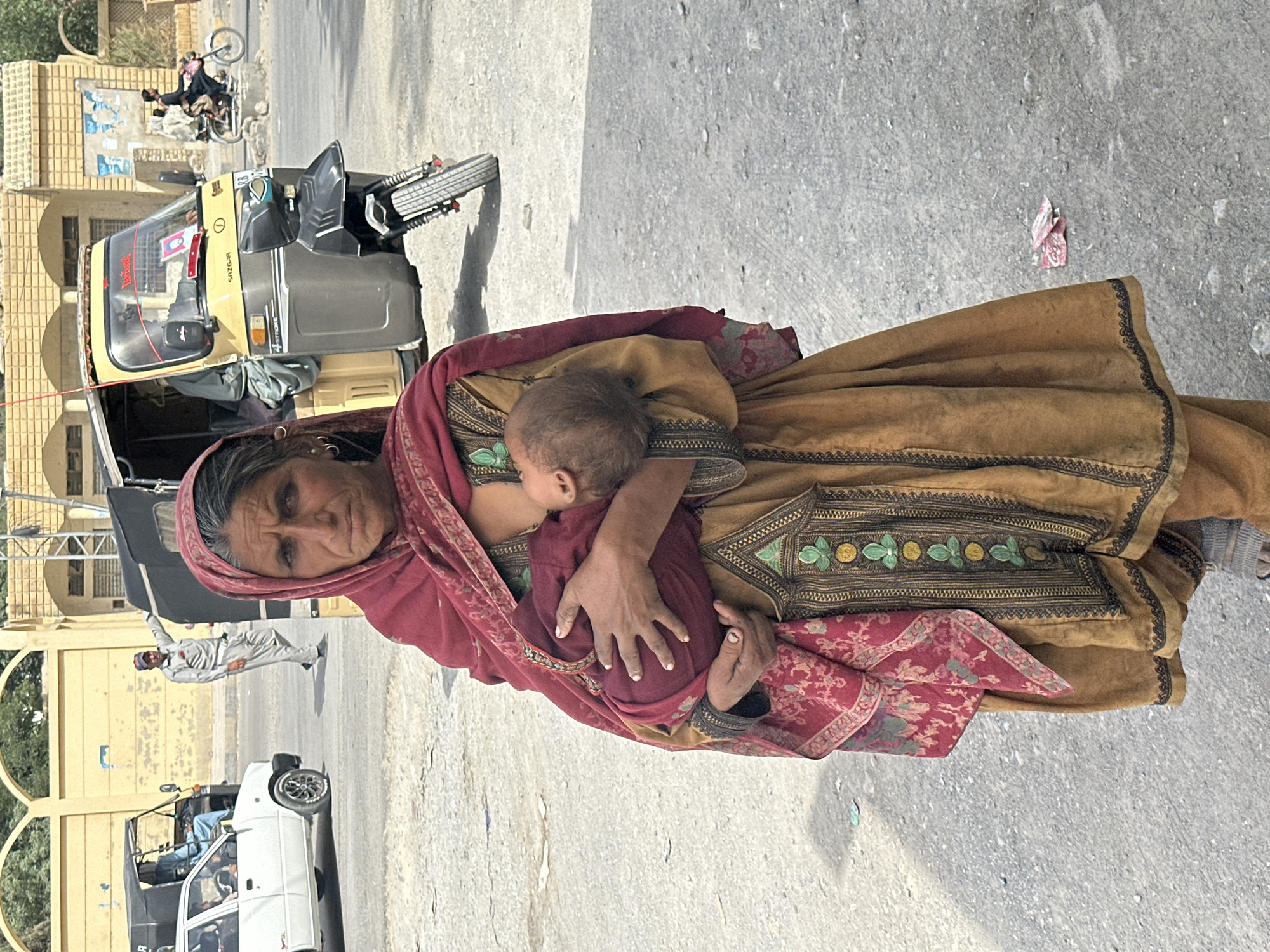In Quetta City Tehsil, Pakistan, a woman stands on a paved surface, holding a baby. She wears a tan, embroidered dress covered with a red shawl. A red scarf is on her head. Several auto rickshaws and a white vehicle are in the background. A man in light-colored clothing walks on the street behind the woman. A building with arched openings is visible in the background.