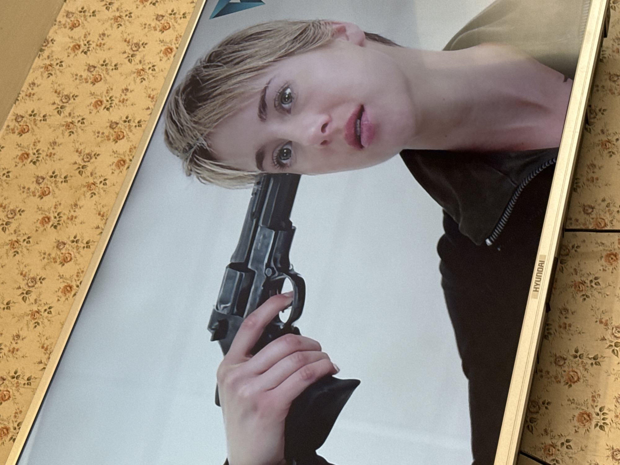 The image shows a framed poster of a woman holding a gun to her head. The poster is leaning against a wall with a floral patterned wallpaper. The frame of the poster has a "HYUNDAI" label on the side. The specific location in Mytishchi, Russia, cannot be determined from the image.
