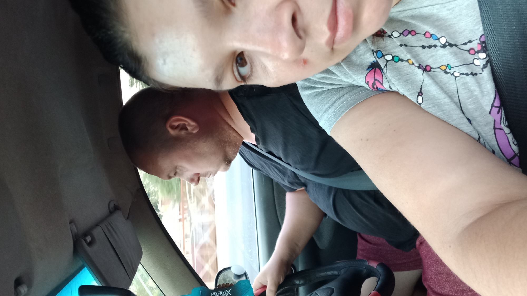 In the image, a man and a woman are in a car. The man is driving and is wearing a black t-shirt. The woman is in the passenger seat and is wearing a grey t-shirt with a colorful beaded necklace design. The car appears to be moving, as indicated by the slight blur of the background outside the windows. The interior of the car shows a sun visor and part of the dashboard. The lighting suggests it is daytime. The photo is likely taken in Kropyvnytskyi, Ukraine, based on the provided context, although no specific landmarks are visible to confirm the location within the city.