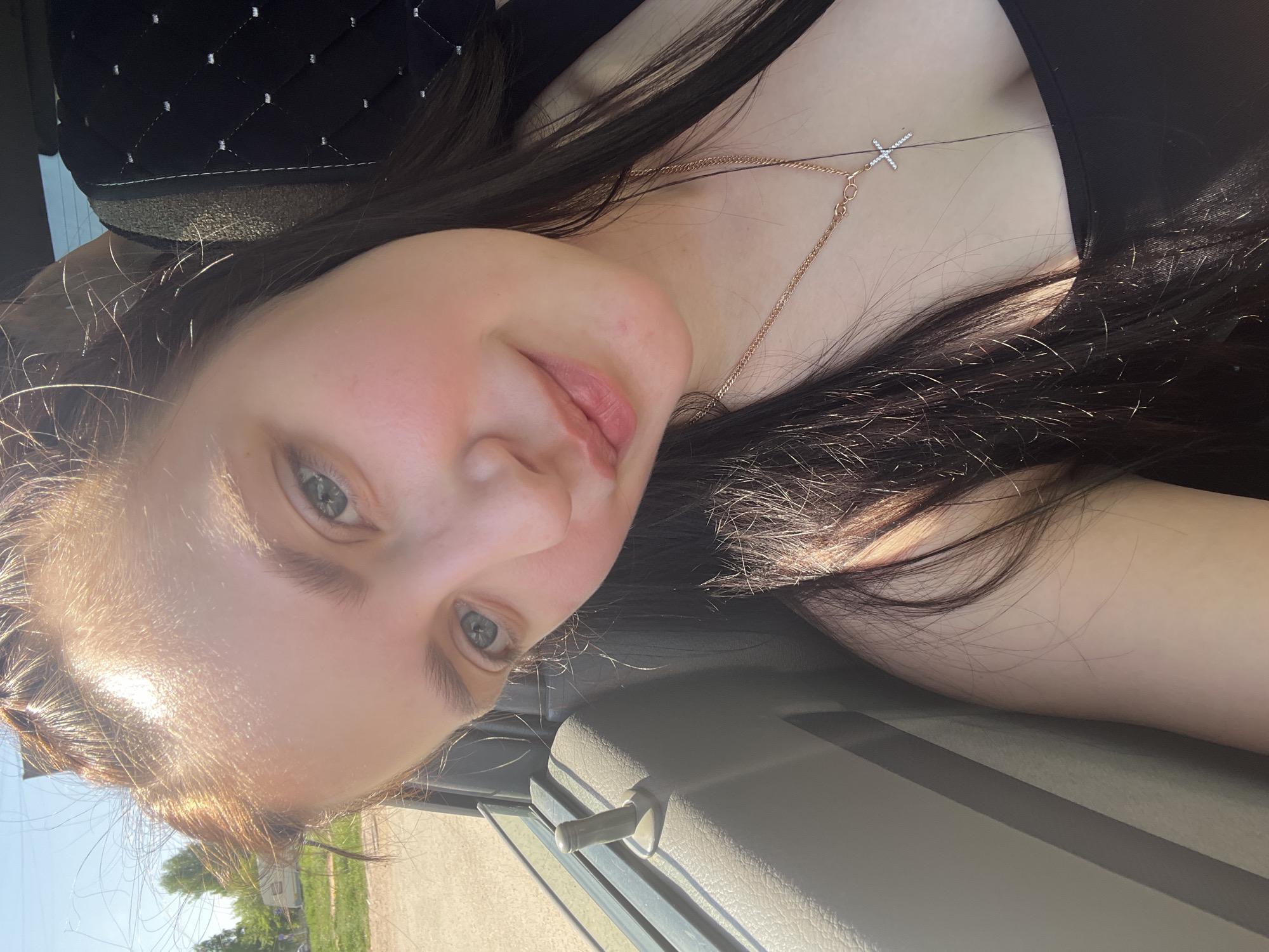 A woman with fair skin and brown hair is visible in this selfie taken inside a vehicle. She is wearing a black top and a gold necklace with a cross pendant. The background shows a residential area, including a house, trees, and a blue sky with bright sunlight. While the prompt requests the location in Chaikovsky, Russia, there is no information to confirm if the photo was taken in that city.