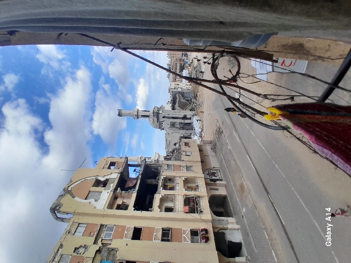 The image depicts a street scene in Khan Yunis, Palestinian Territory, viewed from an elevated perspective, showcasing significant urban destruction. In the foreground on the right, a building wall exhibits entangled utility wires, from which a multi-colored fabric is suspended by a yellow clothespin. Two large blue water bottles and a white sign bearing a red crescent emblem are affixed to this wall. The midground features a two-lane asphalt road with a faded white dividing line, flanked by sidewalks covered in debris. Several individuals are present: one person in dark clothing carries a yellow object while walking on the road, and other smaller figures are discernible further along the street and on the left sidewalk. The left side of the scene is dominated by a multi-story residential building suffering severe structural damage, with large sections of its facade collapsed, revealing exposed interior rooms and numerous broken or missing windows. In the background, a prominent mosque with a tall, slender minaret, largely intact but showing weathering, and a partially damaged dome, stands amidst extensive rubble and collapsed structures. The sky above is partly cloudy, with white cumulus clouds set against a blue expanse.