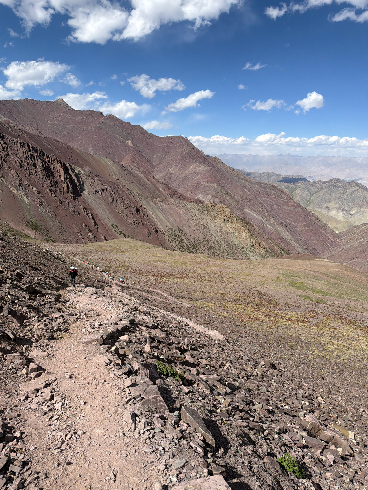 A group of people are hiking on a rocky, mountainous trail under a blue sky with scattered white clouds. The mountains are a reddish-brown color with visible geological layers. The trail is made of dirt and stones, with larger rocks and scree on either side. The vegetation is sparse, consisting of dry grasses and small shrubs. In the distance, more mountains are visible, fading into a hazy blue. The hikers, appearing as small figures in the distance, are carrying backpacks. The scene appears to be in a high-altitude, arid environment.
