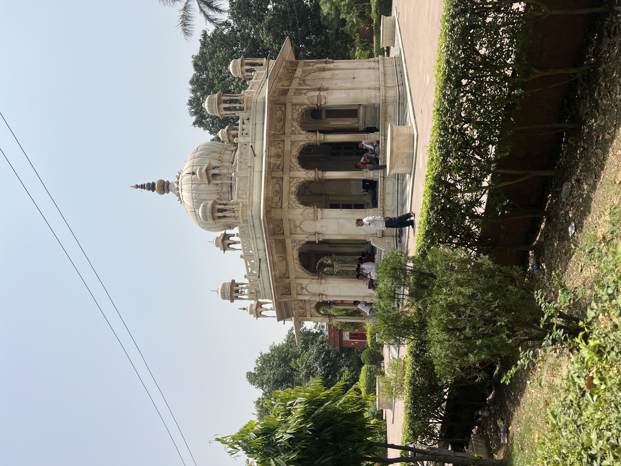 The image shows a white marble structure with a domed roof and arched openings, likely a historical or religious building, situated in a garden or park setting. Several people are present, some inside the structure and one walking nearby. The surroundings include neatly trimmed hedges, trees, and open space. Power lines are visible in the sky. The location is Varanasi, India.