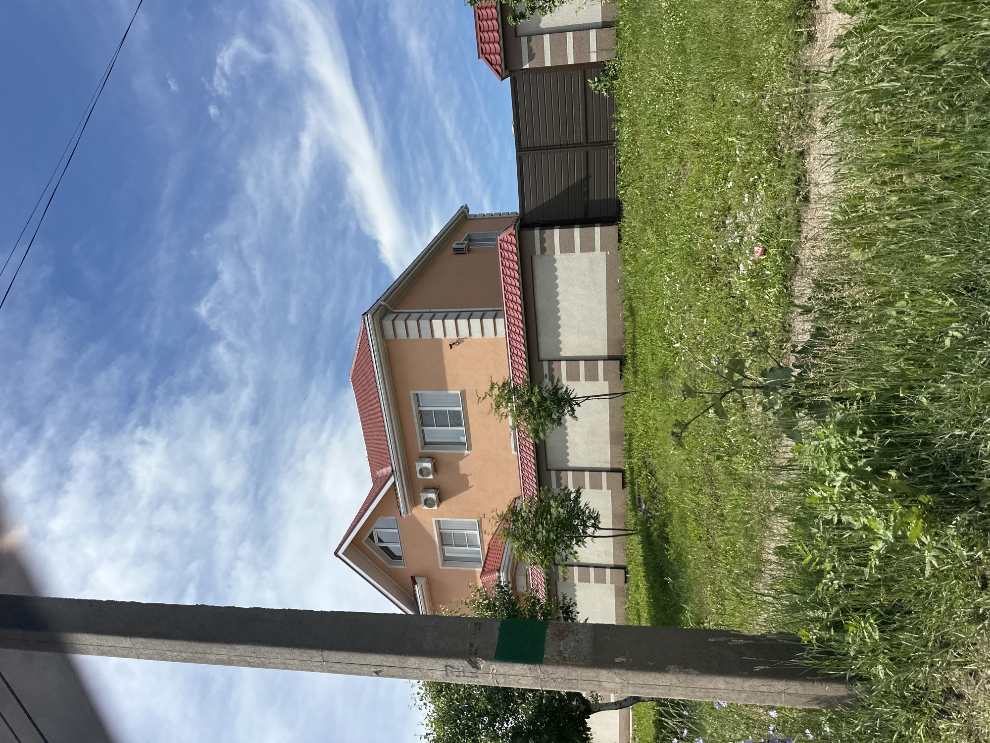 The image shows a two-story house with an orange facade and a red tiled roof, located in the city of Sterlitamak, Russia. The house has several windows, two air conditioning units, and is surrounded by a light-colored fence with a darker brown gate. The foreground is dominated by a concrete utility pole on the left and lush green grass with some wildflowers. The sky is blue with scattered white clouds. The scene appears to be a residential area on a sunny day.