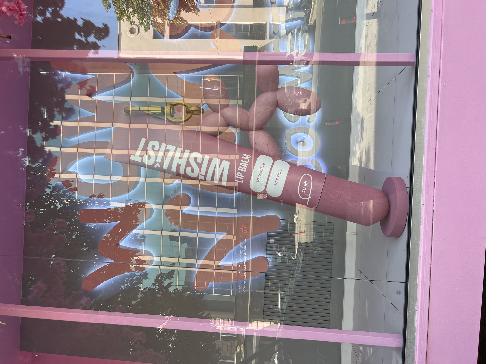 The image shows a storefront display in Odesa, Ukraine. The display features a large, pink lip balm tube with the brand name "WISHLIST" and "LIP BALM" printed on it. The tube is propped up by a stand and has text indicating "VITAMIN E" and "PEPTIDE." Behind the lip balm, a large graphic with glowing blue letters and red cursive text is visible, partially obscured by reflections of trees and buildings. The reflection in the glass shows a street scene with cars and pedestrians. The storefront itself is painted pink.