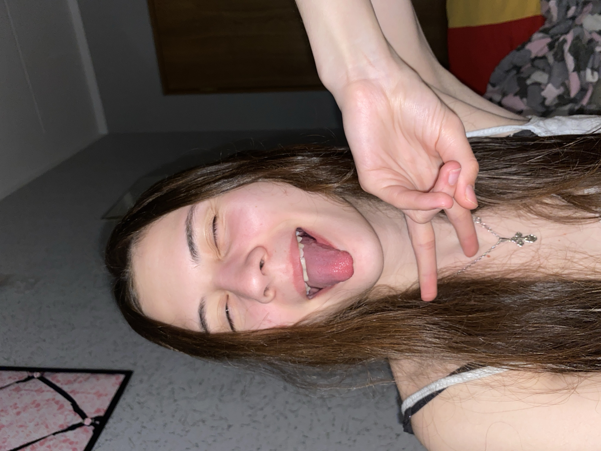 A young woman with long brown hair is lying on a gray floor, sticking her tongue out and making a hand gesture. She is wearing a white tank top with black straps and a silver cross necklace. A portion of a pink and black patterned rug is visible on the floor. A wooden frame is mounted on the wall in the background. A bed with red, yellow, and gray patterned bedding is also partially visible. No definitive evidence suggests the location is Votkinsk, Russia.