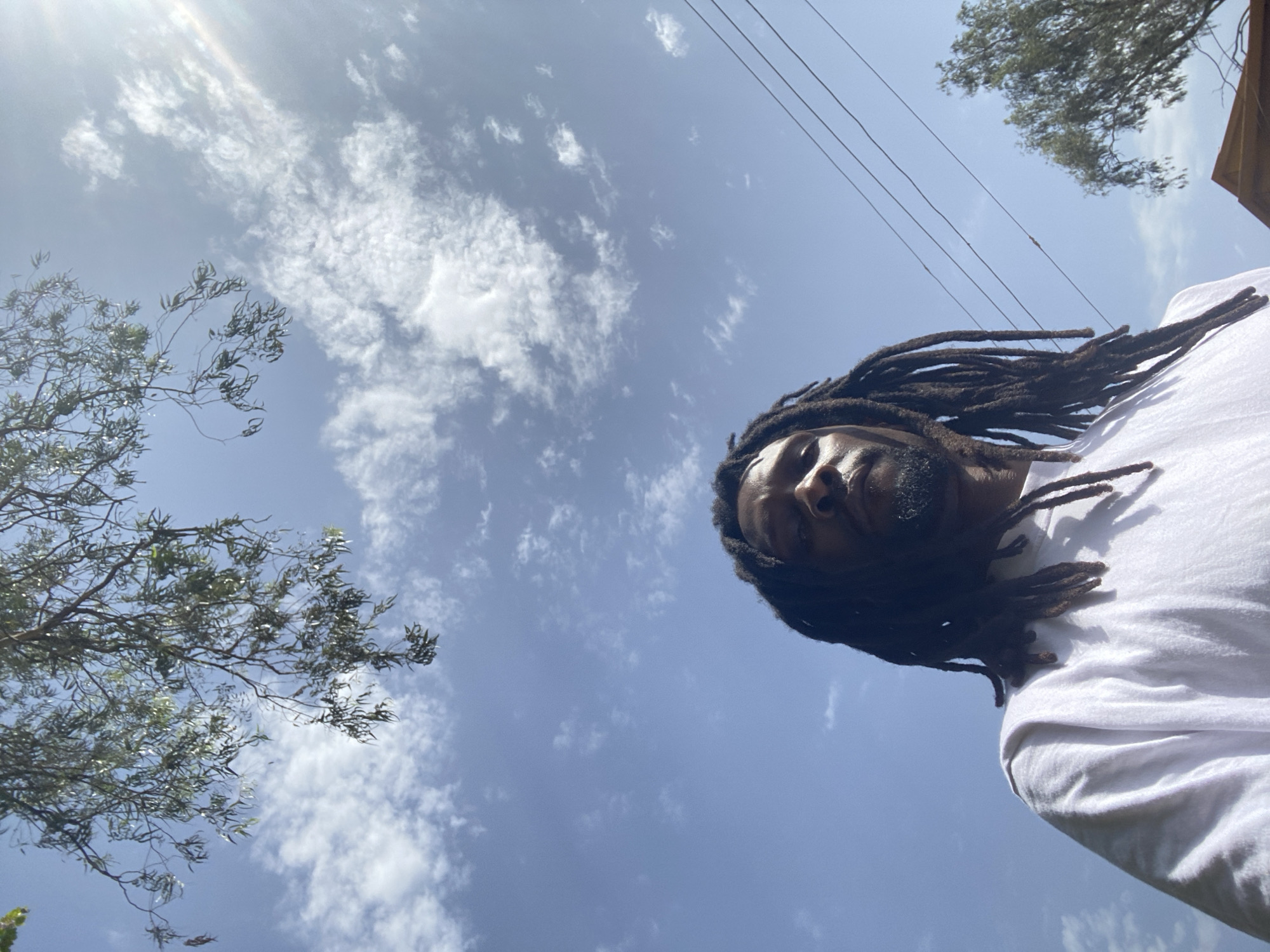 The image shows a low-angle shot of a Black man with dreadlocks, wearing a white t-shirt, looking up at the sky. He is the primary subject, positioned in the lower half of the frame. The background consists of a clear blue sky with scattered white clouds. Sunlight streams in from the upper right, creating a lens flare effect. Tree branches with green leaves are visible in the upper left corner, and some power lines stretch across the sky in the upper right. A portion of a wooden structure is visible in the lower right corner. The scene appears to be outdoors during the daytime. The image does not provide any specific information about the location within Gura Top, Nigeria.