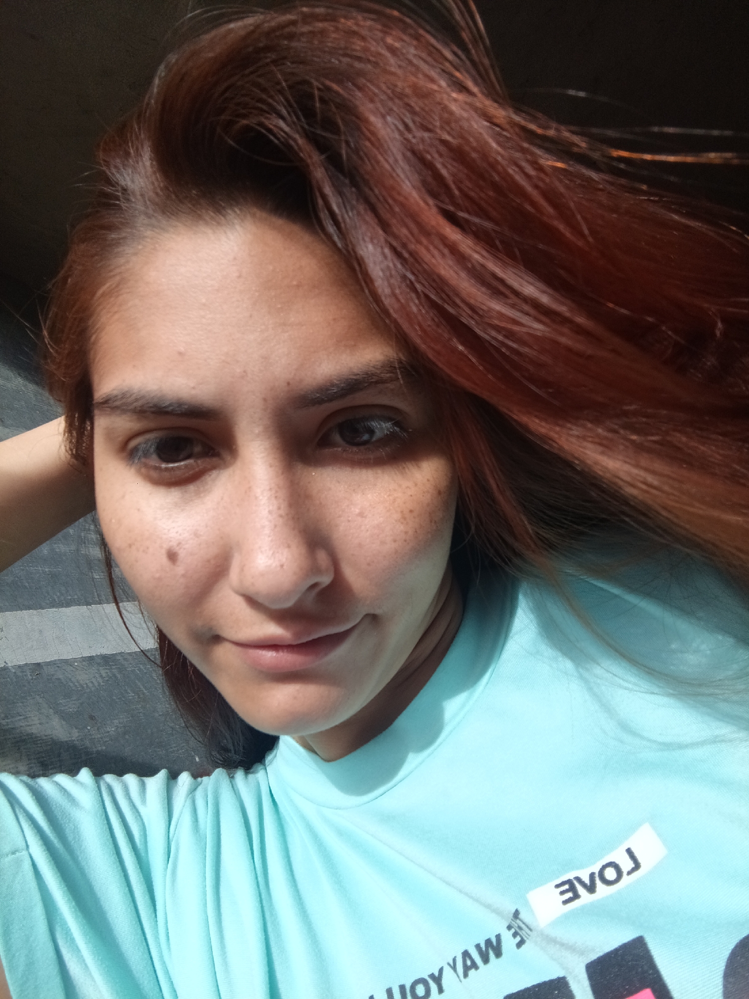 A medium close-up shot shows a woman with auburn hair and light skin wearing a light blue t-shirt with a partially visible word "LOVE" printed on it. Her hair is styled with a side part, and strands of hair fall across her forehead. She has visible freckles on her face and a mole on her left cheek. The background shows a gray, slightly textured surface. The picture was taken in Juan Griego, Venezuela.