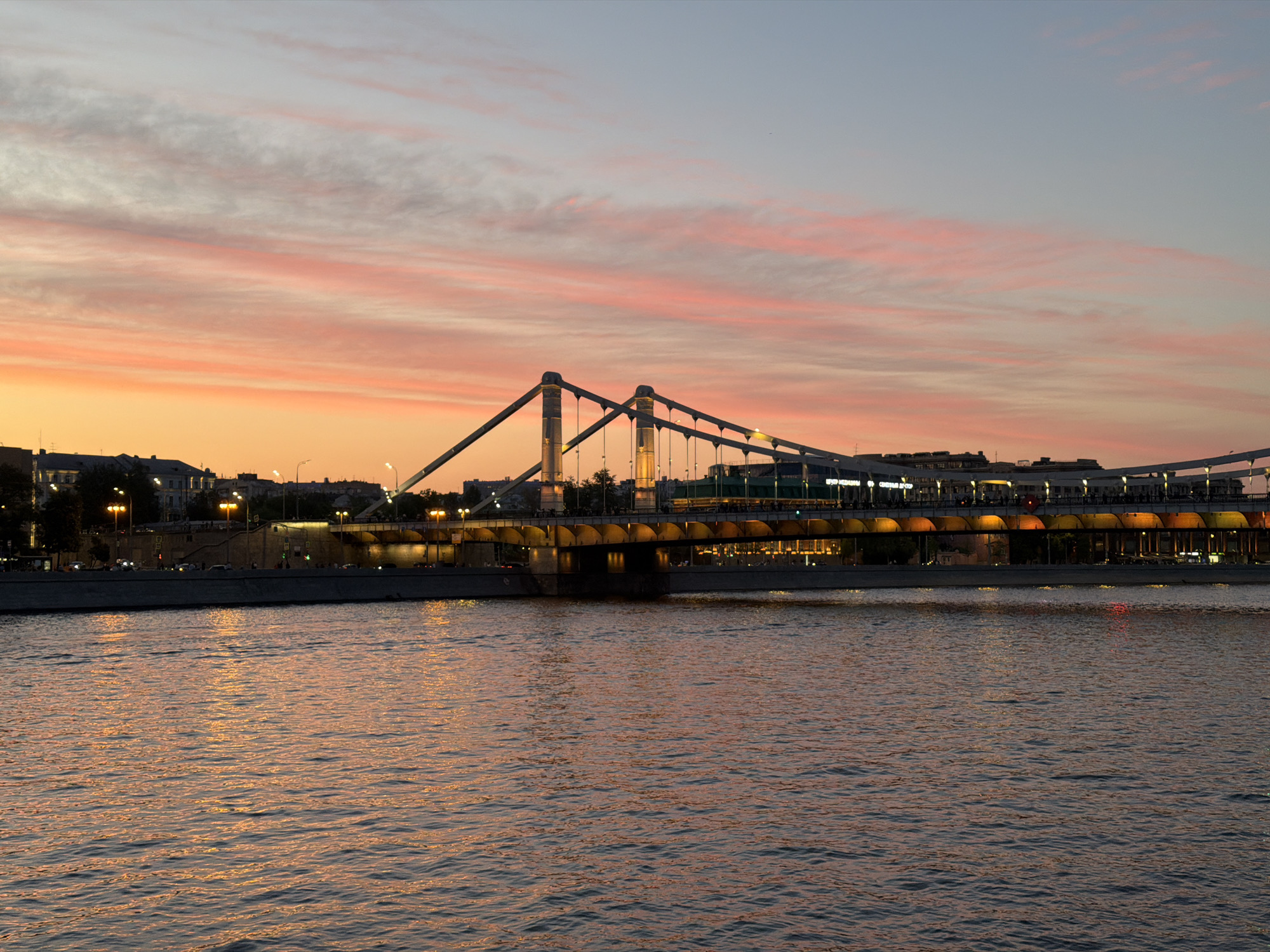 The image captures a cityscape at twilight, featuring a prominent suspension bridge over a body of water. The bridge is characterized by its unique "M" shaped pylons. Buildings line the opposite bank, illuminated by streetlights. The sky above is a mix of pastel hues, with orange and pink streaks hinting at the fading sunset. The water reflects the ambient light, creating a shimmering effect. The general location appears to be in Moscow, Russia, likely depicting the Krymsky Bridge.