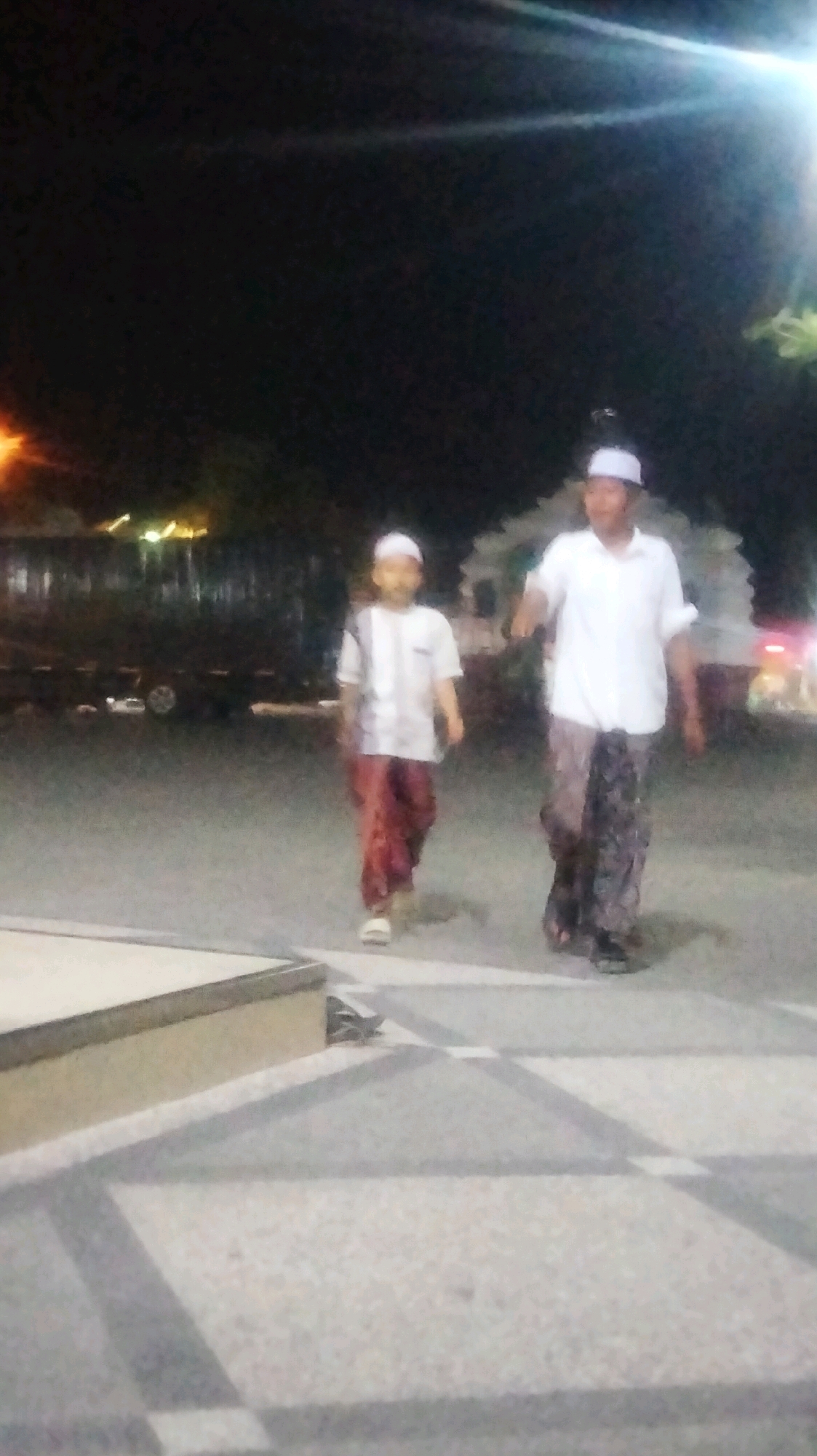In the city of Soko, Indonesia, two individuals are captured in a nighttime scene. The image, taken at night, shows a young boy and an adult male walking. The boy, on the left, is wearing a white striped shirt and red pants, with a white cap. The adult male, to the right, is dressed in a white short-sleeved shirt and patterned sarong, also wearing a white cap. They appear to be walking on a paved area with some patterned squares. In the background, blurry lights suggest vehicles and possibly buildings or trees. The overall atmosphere is dark, with artificial lighting creating some glare and highlights in the image.