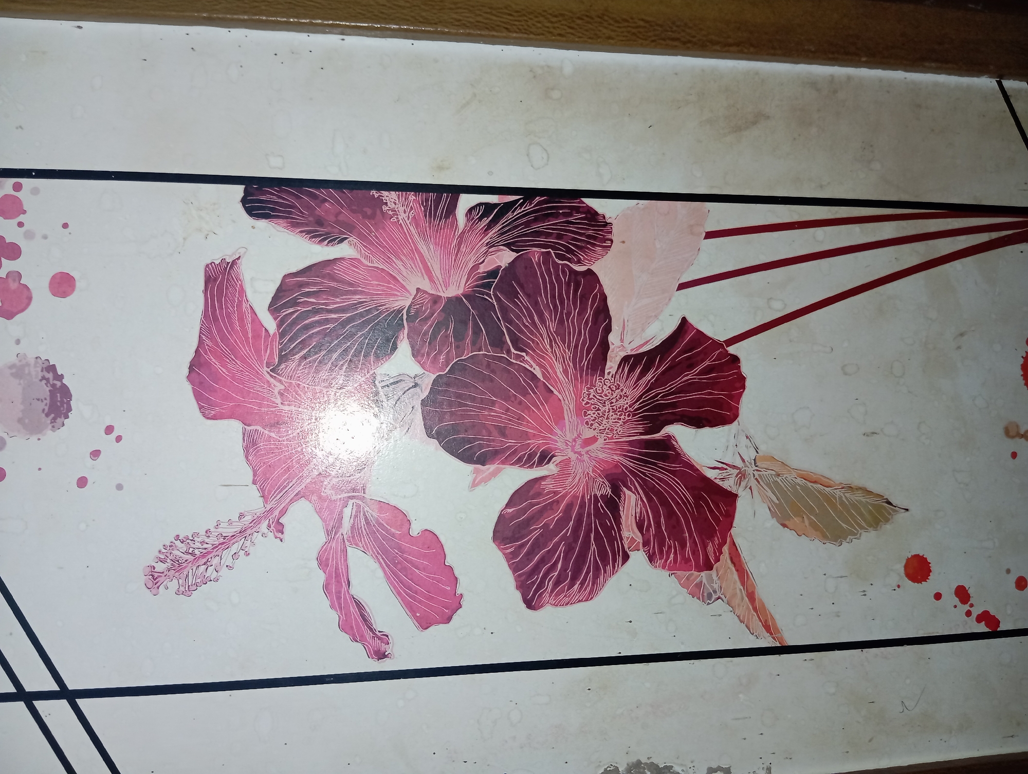 A close-up, vertical, eye-level shot shows a decorative panel featuring hibiscus flowers. The panel is white with a textured, slightly mottled surface and is bordered by thin black lines on the left and a brown textured surface on the right. The hibiscus flowers are depicted in shades of pink and deep red, with white line art highlighting the veins of the petals and the stamens. Some petals are in sharp focus, while others are slightly blurred. Red and pink splatters, resembling paint or ink, are scattered across the white background, particularly near the bottom. The overall impression is of a stylized floral design. The location is the city of Rejosari, Indonesia.