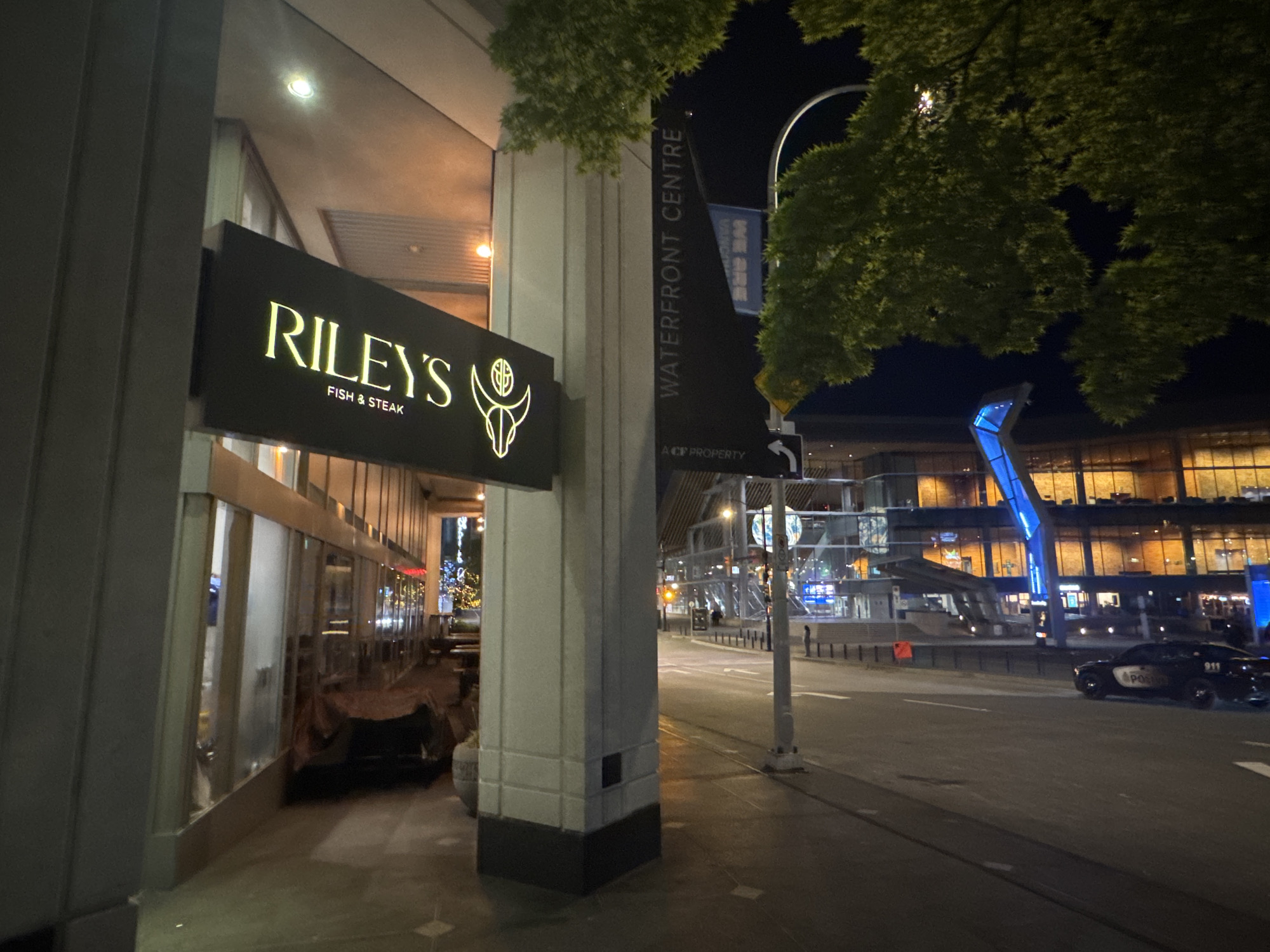 The image depicts a street scene at night in Vancouver, Canada, specifically at the Waterfront Centre. The focus is on "Riley's Fish & Steak" restaurant, indicated by a large sign. A black banner for "Waterfront Centre" is also visible. The street is mostly empty, save for a black police vehicle parked on the right side. In the background, a building is illuminated with warm interior lights and a structure is highlighted with blue lighting. Trees are present in the upper part of the image, partially obscuring the night sky.