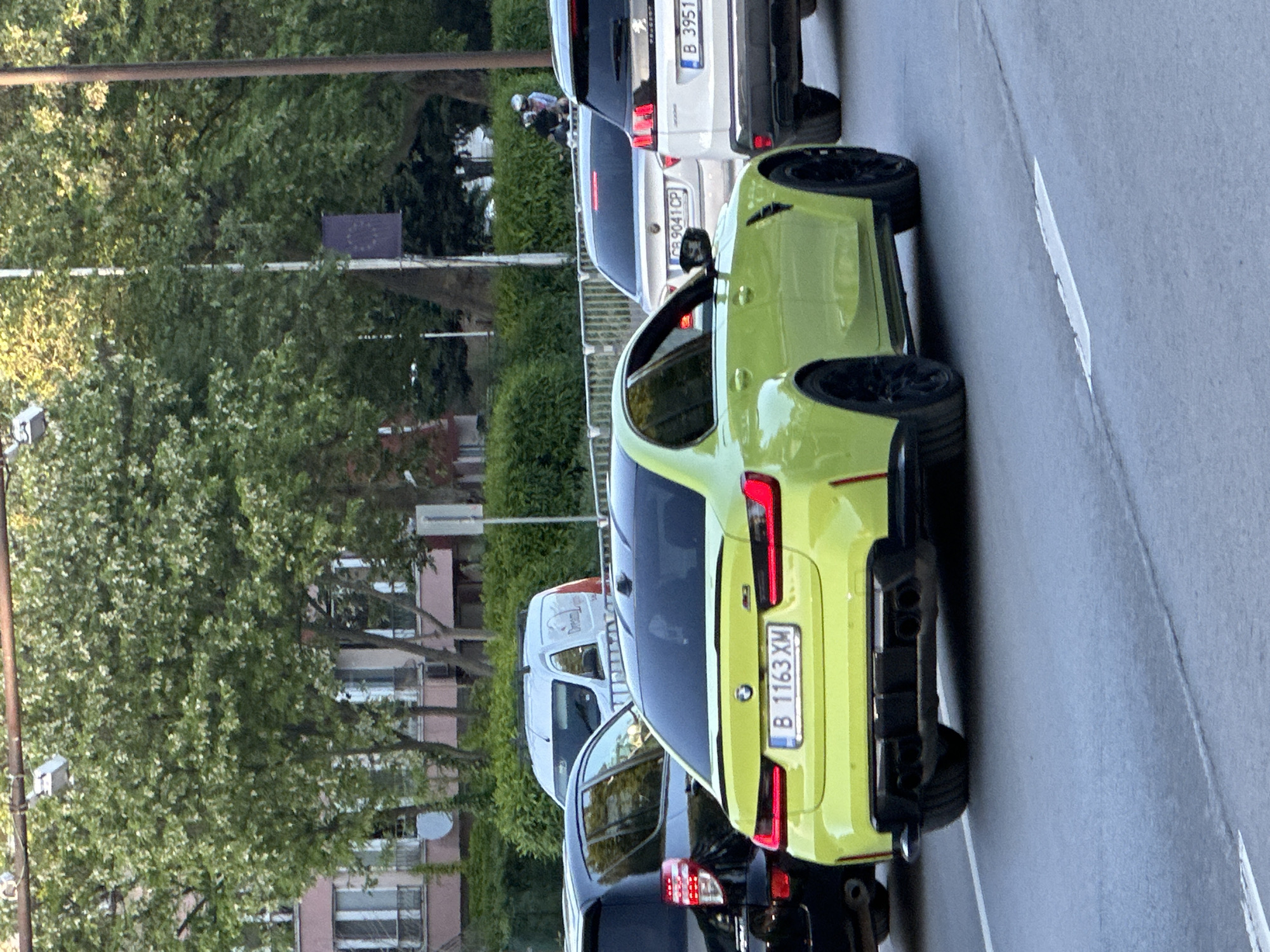 The image shows a street scene in Varna, Bulgaria. A line of parked vehicles is visible along the roadside. A chartreuse BMW is parked in the line of vehicles, with its license plate B 1163 XM clearly displayed. Additional parked vehicles, including a white van and a dark-colored SUV, are also visible in the frame. In the background, buildings and greenery are visible, suggesting an urban setting. Two cyclists are visible between parked cars.