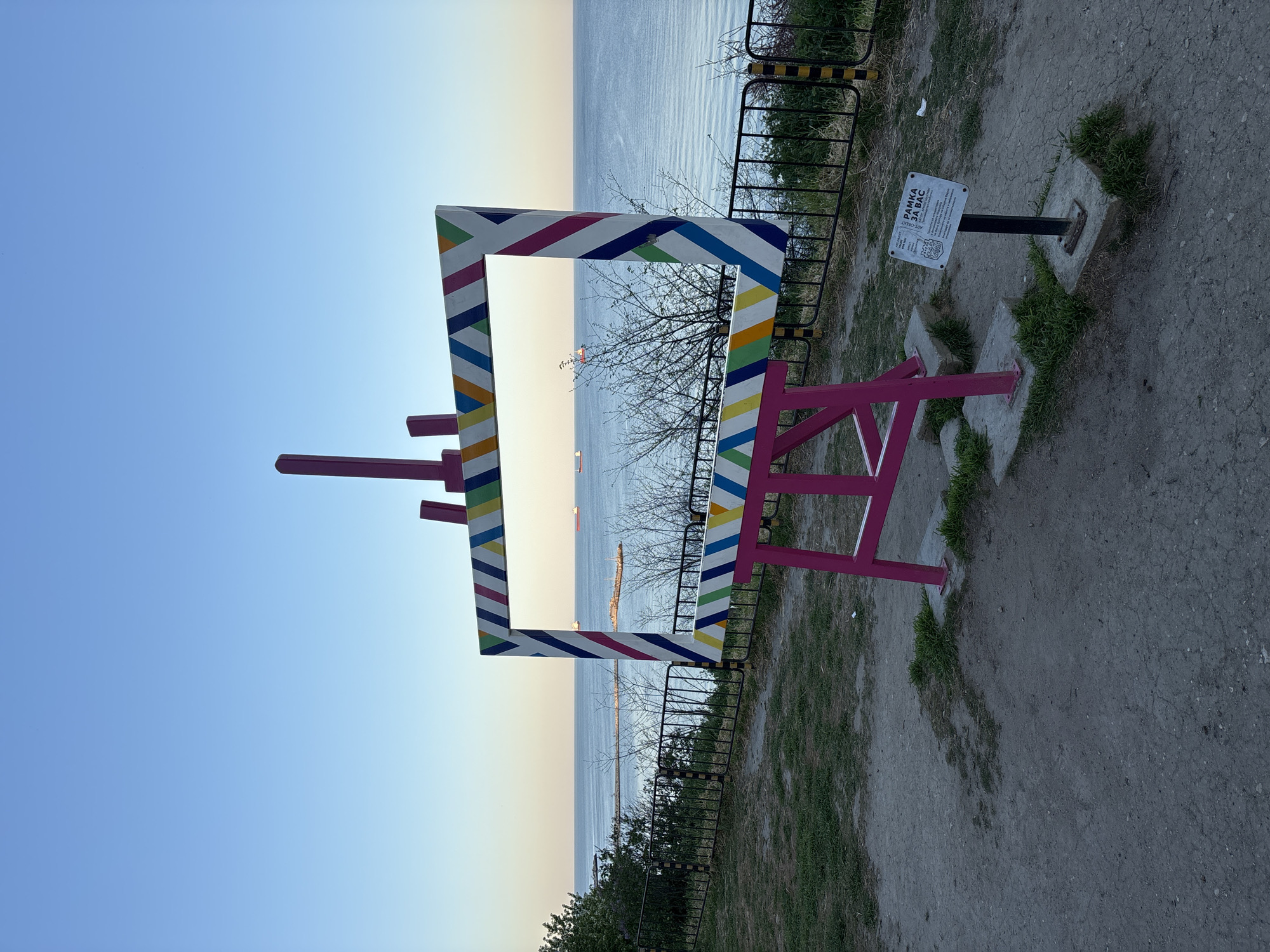 The image shows a colorful, easel-like frame set up as a photo opportunity, looking out over the Black Sea in Varna, Bulgaria. The frame is painted pink with a white border and diagonal stripes of blue, yellow, and green. It has two pink "legs" holding it up and two "brushes" attached to the left side. The scene includes the sea in the background, a low black fence with yellow accents, and a small sign on a post to the right. The ground is a mix of dirt and grass. The sky is clear and light blue. Two ships can be seen in the distance in the sea in the middle of the frame.