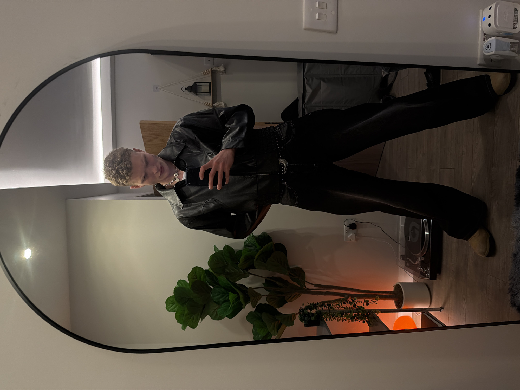 A young man is captured in a full-body selfie taken in a large, arched mirror with a black frame. He is standing upright, holding a black smartphone in his left hand, angled slightly downwards as he takes the photo. He has short, curly light-blonde hair and fair skin.
He is dressed in an all-dark outfit, consisting of a black leather jacket with a collared neckline and pockets, dark trousers, and light-colored shoes, possibly tan or cream, peeking out from beneath his trousers. A silver chain necklace is visible around his neck. His expression is neutral or slightly smiling.
The reflection in the mirror reveals a room with light-colored walls and a light wooden floor. In the background, behind the man, there's a light switch with two white toggles on the wall, and a decorative macrame hanging with a small black lantern. Further to the right, two white electronic devices, one with a blue indicator light, are plugged into a wall outlet.
In the lower part of the reflection, a large artificial fiddle-leaf fig plant in a white pot stands on the floor, next to a smaller plant. An orange glowing lamp or orb is also visible on the floor, casting a warm light. Next to the plants, a turntable/record player is placed on a low stand, with a grey rug partially visible on the floor beside it. The indoor lighting suggests the photo was taken in the evening or at night. The setting appears to be a modern indoor space, likely in London, United Kingdom. No visible text is discernible.