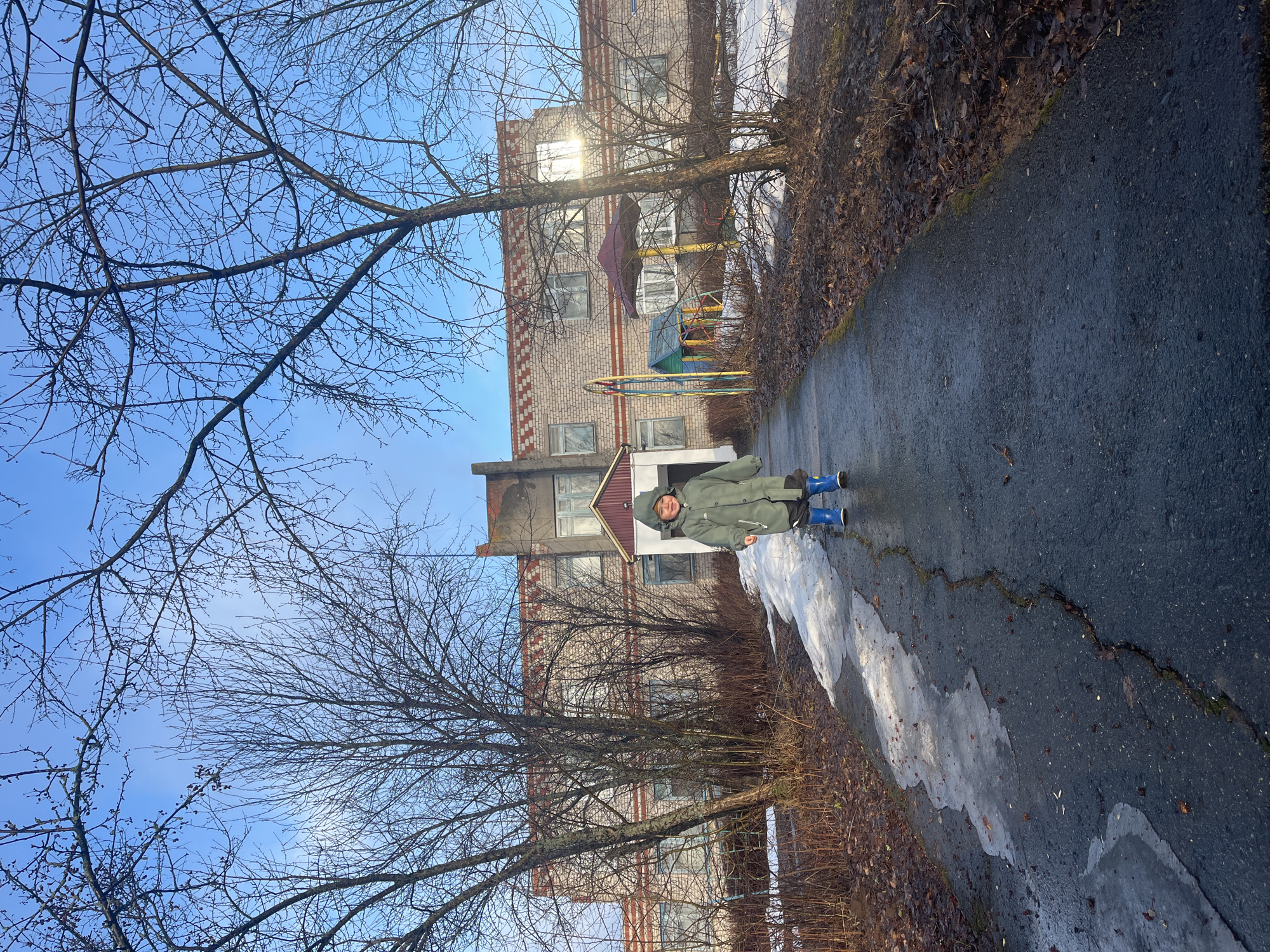 A person in a green coat and blue boots stands on a paved path in Pavlovka, Russia. The path has patches of ice and is bordered by leafless bushes and trees. A brick building with a playground is in the background. The sky is blue with bare tree branches visible.